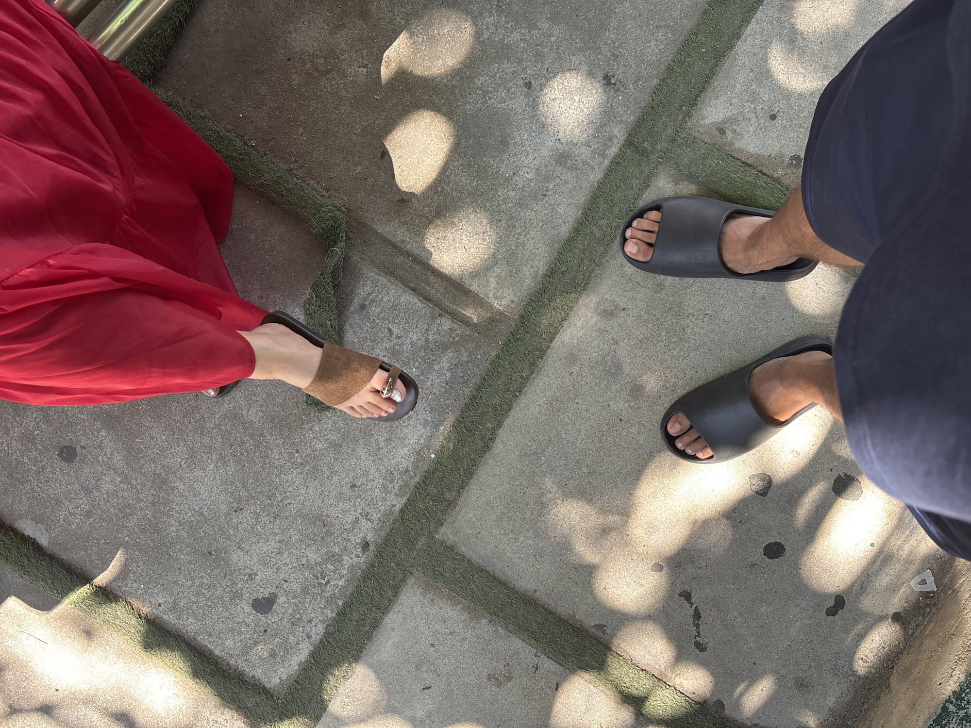 Two people's feet are visible from a high angle perspective, standing on a concrete surface with green bordered lines. The person on the right wears a red dress and brown sandals, with their left foot showing. The person on the left wears dark blue shorts and black slides, with their feet fully visible. Sunlight casts dappled shadows on the ground.