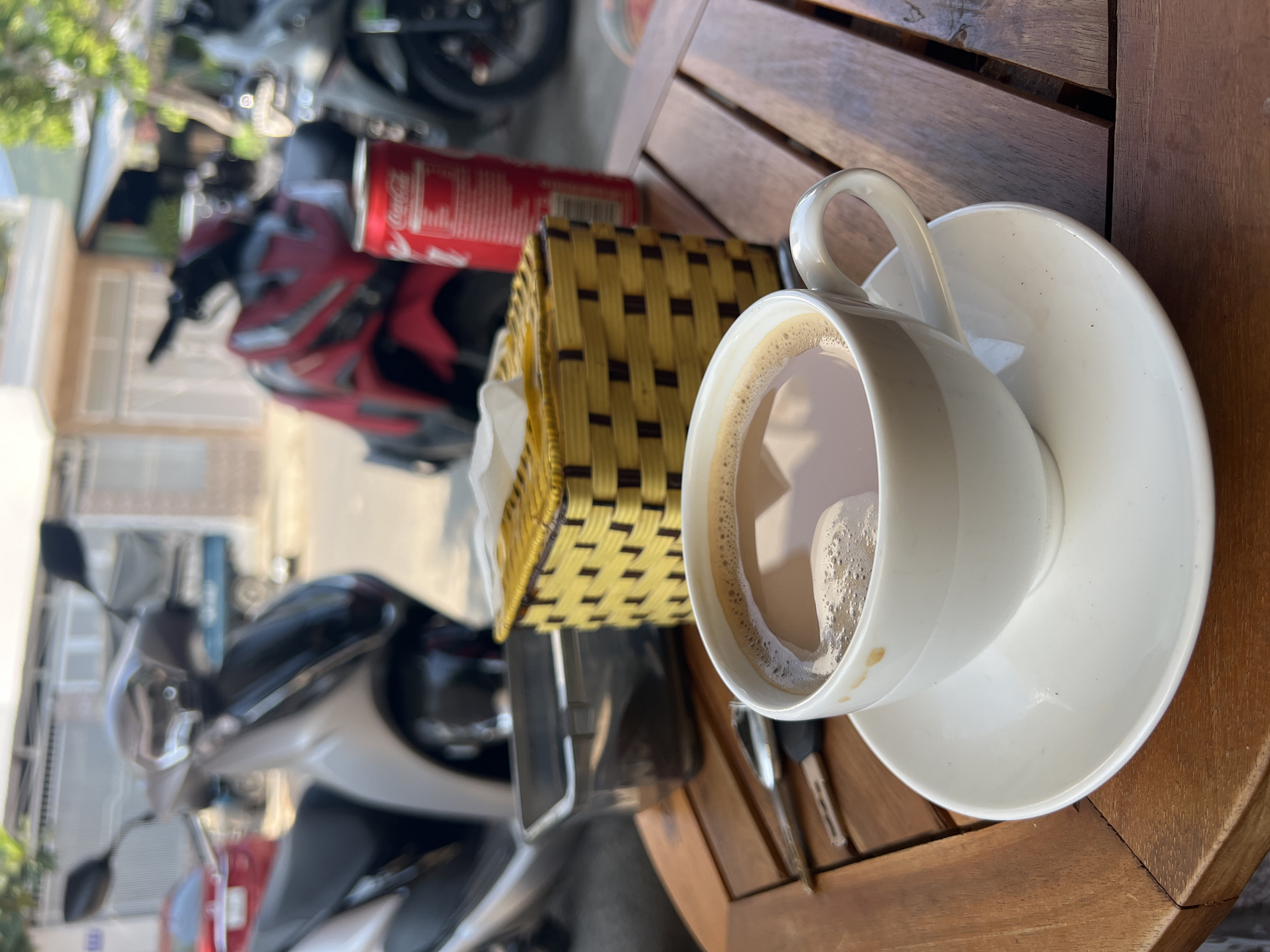 A cup of coffee on a saucer sits on a wooden table outdoors. A yellow woven basket, a red soda can, and several motorbikes are visible in the background. The location is likely in Bắc Nha Trang, Vietnam.