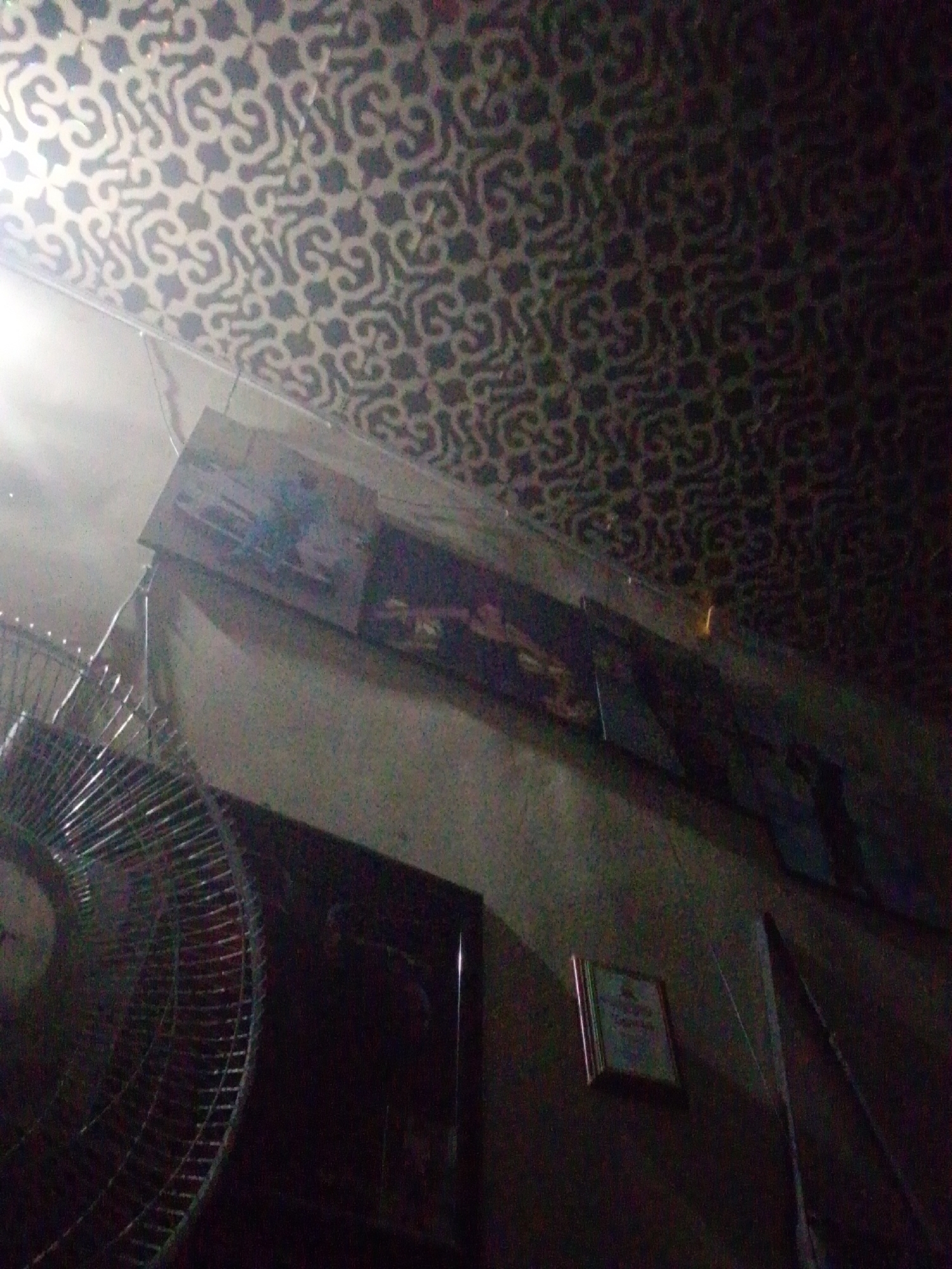 The image depicts the interior of a dimly lit room in Unknown, Unknown, taken from a low-angle perspective looking upwards. The ceiling features a repeating pattern of dark, stylized curvilinear motifs against a lighter background; the upper left portion of the ceiling is brightly illuminated by an unseen light source, creating a strong glare. The walls below the ceiling are a light, neutral color, appearing largely in shadow towards the right side of the frame.
In the lower-left quadrant, a large, metallic floor or pedestal fan, characterized by its circular protective grille, is prominently positioned. Above the fan, on the wall, several rectangular objects are horizontally aligned; these appear to be framed pictures or decorative panels, with their specific contents indistinct due to the low light and distance. A smaller, square-framed item is visible further to the right on the wall, below the line of larger frames. Thin electrical wires or cables are observed hanging down from the brightly lit area in the upper left and also beneath the horizontal frames. The overall scene is characterized by deep shadows and visual noise, suggesting low-light photography. No individuals or specific actions are discernible.