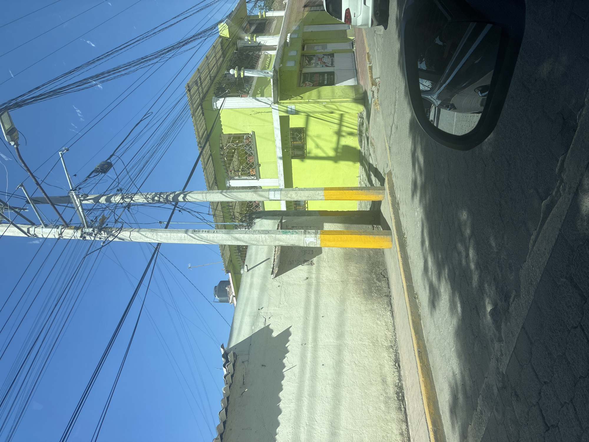 The image shows a street scene in Cuautitlán Izcalli, Mexico, featuring a utility pole laden with numerous electrical wires against a clear blue sky. A two-story building painted a bright green color stands adjacent to the utility pole and the street. The building features barred windows and signage indicating that it might be a commercial establishment. The image is framed from the perspective of a vehicle, with a car side mirror visible in the upper right corner, obscuring part of the street. The road surface appears to be made of asphalt. Sidewalks with yellow safety stripes are along the edge of the street. The lighting indicates it is daytime with sunlight casting shadows on the buildings and pavement.
