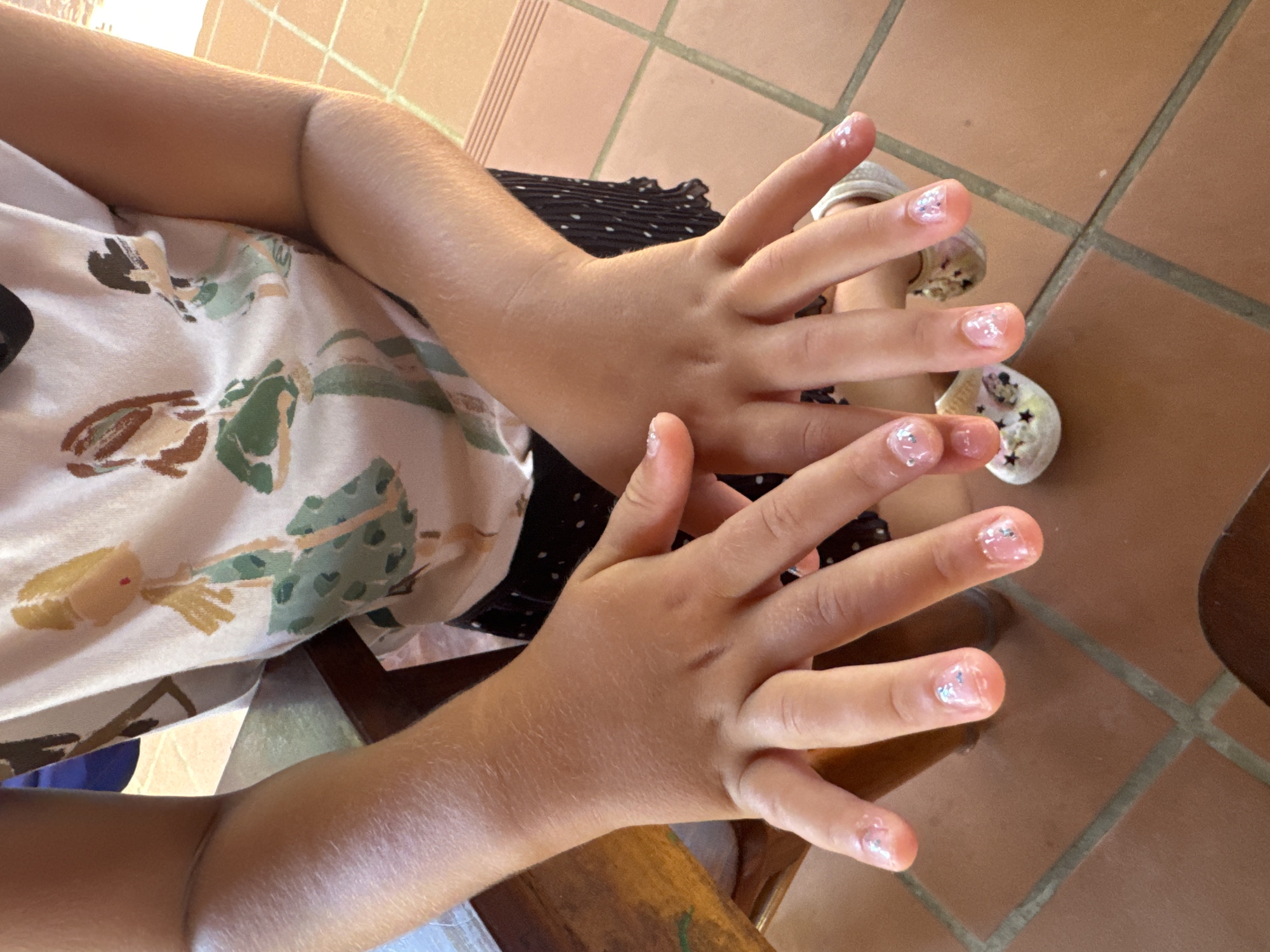 A child's hands are held up, displaying clear nail polish with silver glitter. The child is wearing a patterned shirt and a black and white polka dot skirt. The background shows a tiled floor. The location is not possible to determine from the image.