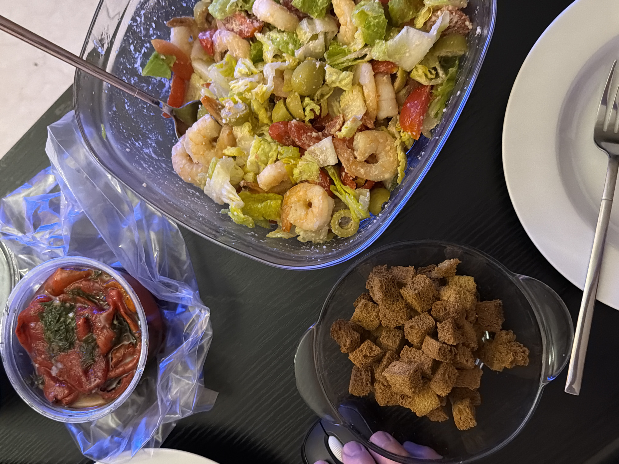 An overhead shot captures a casual meal set on a dark table. The primary subject is a large, clear glass bowl brimming with a shrimp salad, consisting of cooked shrimp, chopped green lettuce, red tomato pieces, and green olives. A light-colored dressing or powdered seasoning is visible on the salad ingredients, and a silver serving spoon rests within the bowl.
To the bottom left, a small, clear plastic container holds roasted red peppers, garnished with green herbs, likely dill, and is enclosed in a clear plastic bag. Adjacent to this, in the bottom right, a smaller clear glass bowl is filled with square, brown croutons, and a hand with light-colored fingernails is seen holding this bowl. A white ceramic plate and a silver fork are positioned to the right of the main salad bowl, indicating a place setting for a diner.
The image portrays an indoor setting, likely a home, with a meal prepared for consumption. There are no people visible, only a hand. No text is visible in the image. The setting suggests a relaxed dining experience. This meal is located in Tel-Aviv, Israel.