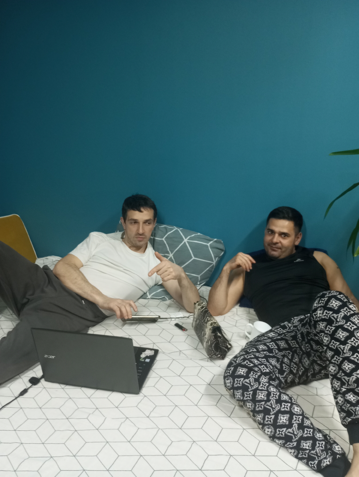 Two men are lying on a bed in what appears to be an indoor residential setting. The man on the left is wearing a white t-shirt and gray pants, pointing towards a tablet or phone that he is holding in his hand. A laptop is also visible near him on the bed. The man on the right is wearing a black tank top and patterned black and white pajama pants. A white mug is next to him on the bed. The bedding has a geometric pattern. The wall behind them is painted a teal color. A plant is partially visible in the top right corner. There is no contextual evidence to indicate the location is in the city of Pervomaysky, Russia. The objects, actions, interactions, and environment are consistent with a scene of relaxation, watching something on a laptop, or engaging in discussion.
