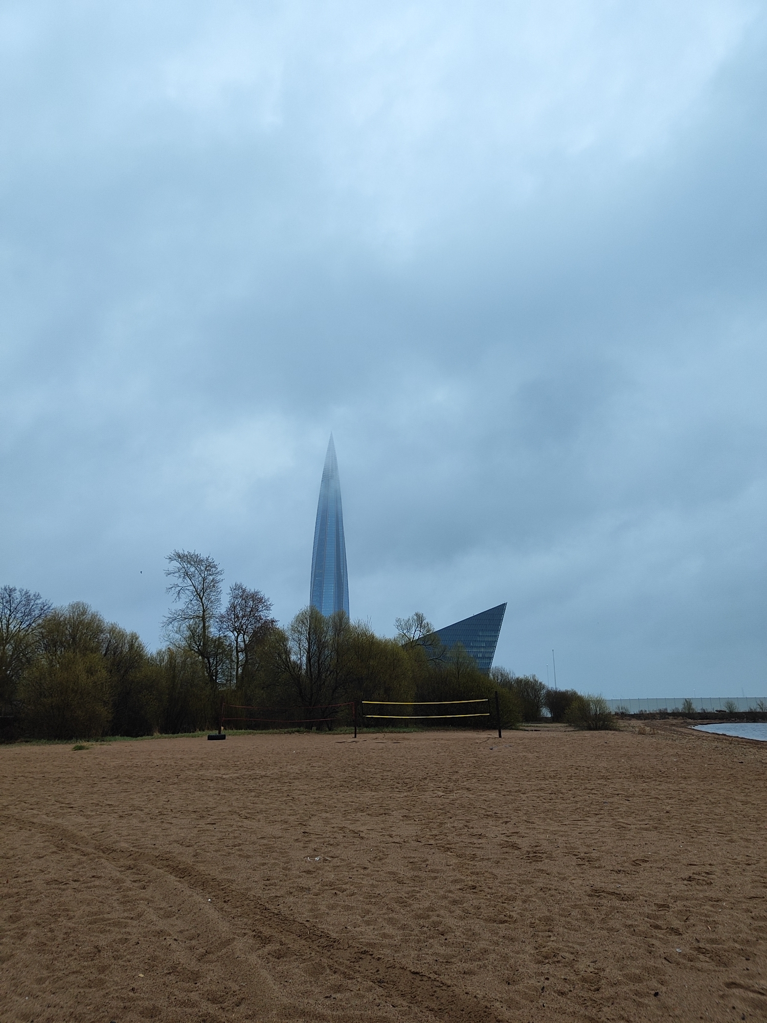 The image depicts a beach volleyball court on a sandy beach. In the background, obscured by clouds, is the Lakhta Center, a skyscraper located in Saint Petersburg, Russia. The sky is overcast. There are some trees and bushes between the beach and the buildings. The volleyball net is in the middle of the scene, in front of the Lakhta Center. Tire tracks are visible on the sand.