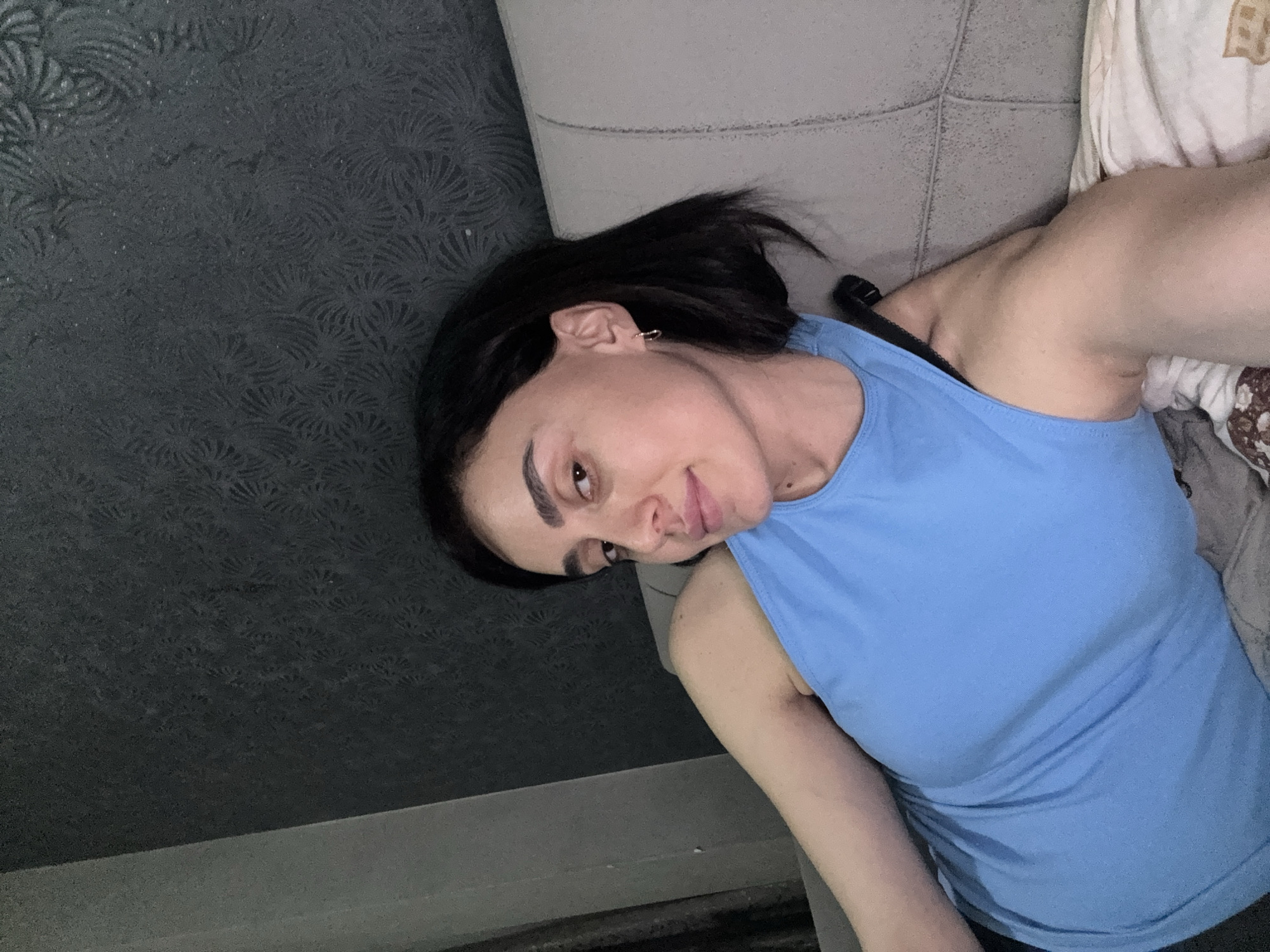 A woman with dark hair is reclining on a couch. She is wearing a blue tank top. The background includes a textured wall in a dark shade and part of the couch. The location is likely an interior setting, though no other details related to Tbilisi, Georgia are present.