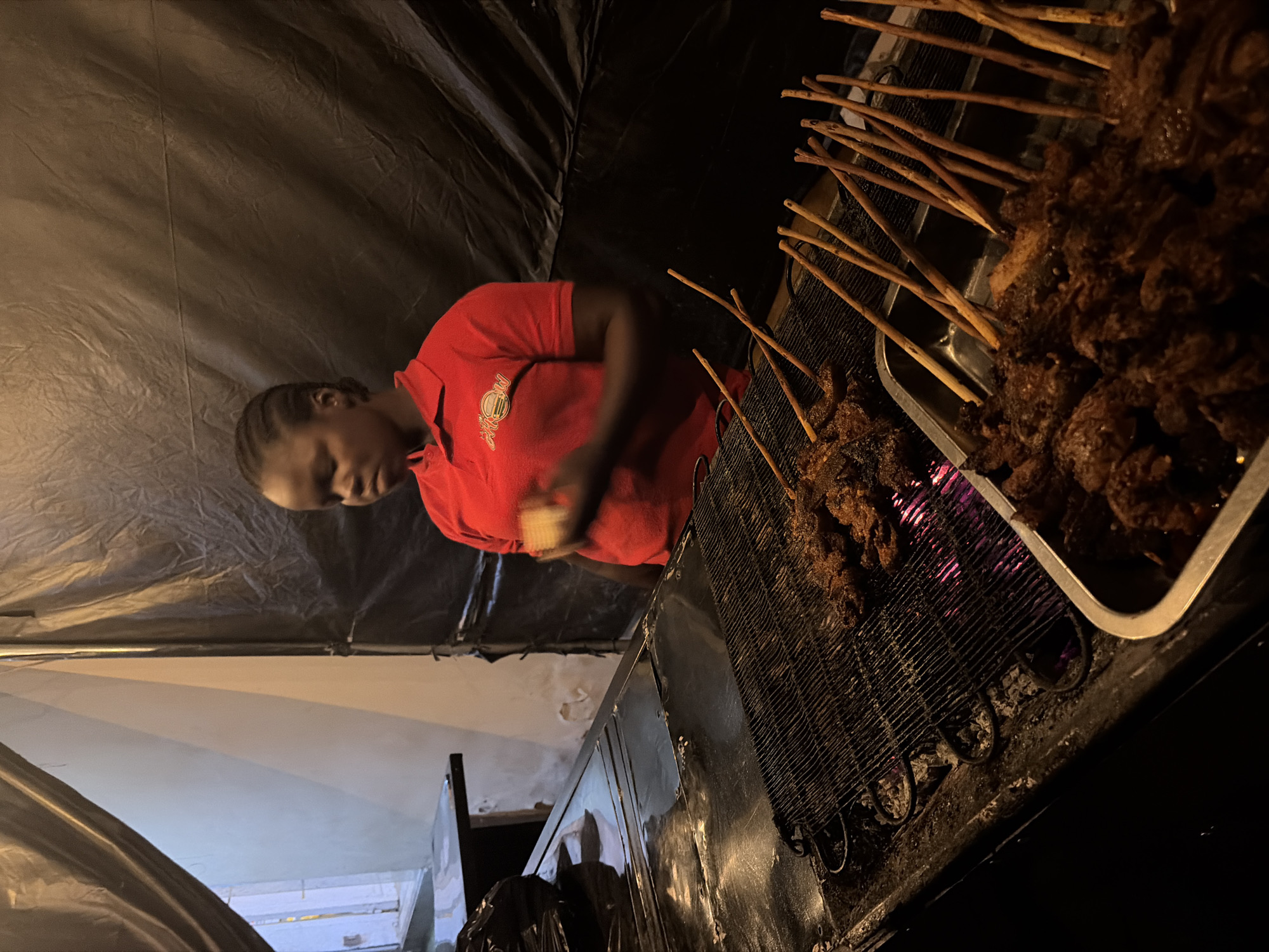 A woman in a red polo shirt is standing behind a counter with a grill in front of her. She has her right hand raised and is looking down. The grill is filled with meat on skewers, and some of the meat is already cooked. In front of the grill is a metal tray also containing meat on skewers. The background is a black tarp. The scene appears to be an outdoor food stall, possibly in the city of Jos, Nigeria.