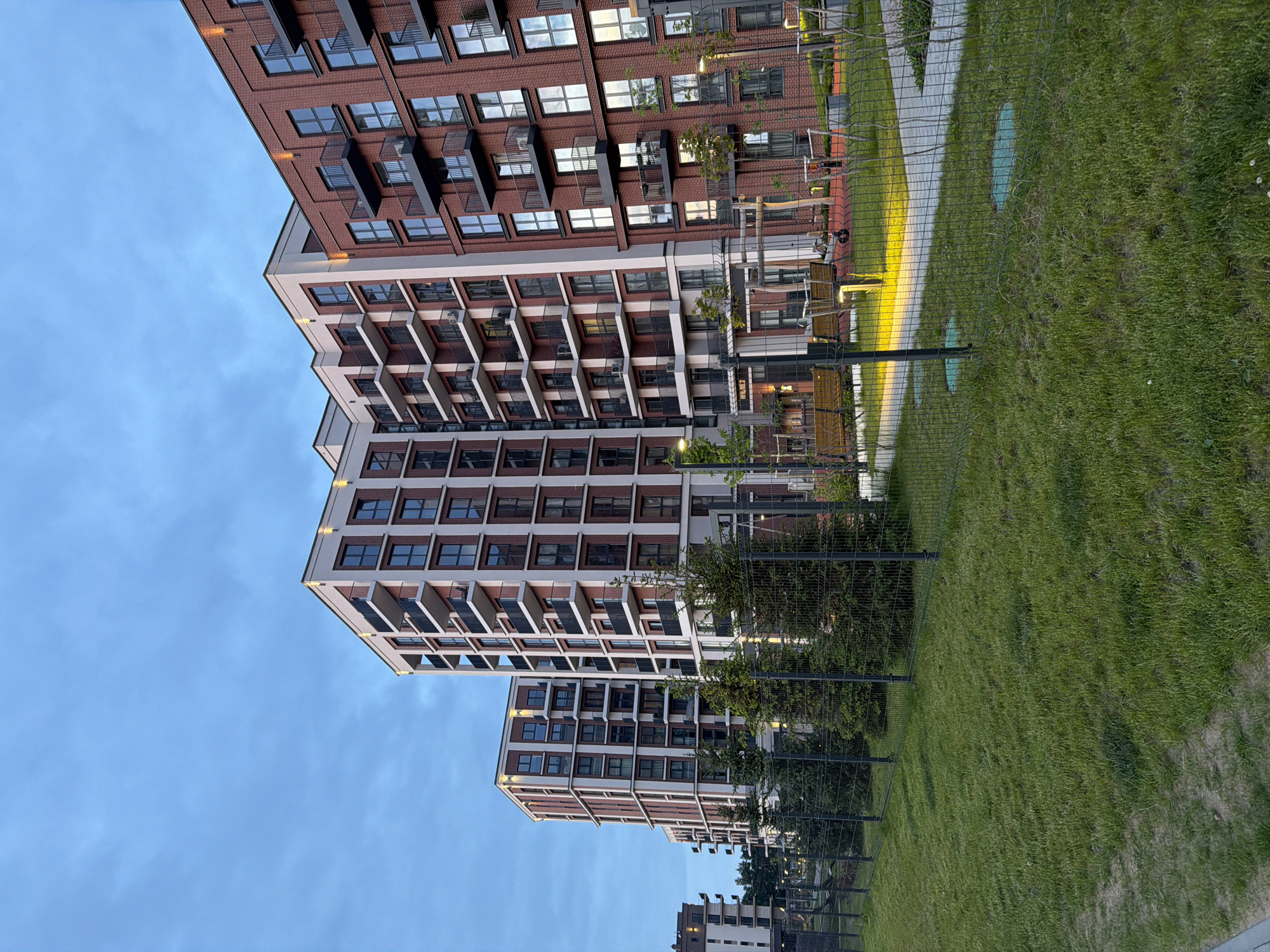 The image captures a modern residential complex in Minsk, Belarus, during dusk or early evening. The scene is dominated by several multi-story apartment buildings exhibiting contemporary architecture. The building on the far right features a prominent red brick facade with numerous dark-framed windows and dark metal balconies. Adjacent buildings to its left present a mixed facade of light-colored (likely white or cream) panels alternating with sections of red brick, also incorporating dark-framed windows and balconies. Artificial warm-toned lighting fixtures illuminate the upper portions of the buildings and ground pathways.
In the foreground, a strip of green grass is visible, bordered by a dark-colored wire mesh fence that runs horizontally across the midground. Behind the fence, a row of small to medium-sized evergreen trees is planted. Further beyond the fence and trees, a paved pedestrian walkway is illuminated by ground-level light fixtures casting yellow light. Two wooden benches are positioned near the walkway. A section of a playground with a red safety surface and some equipment is partially visible in the mid-right. Three green circular manhole covers are embedded in the grass near the fence. The sky above is a pale blue with faint cloud cover, consistent with twilight conditions. No individuals are discernible within the frame.