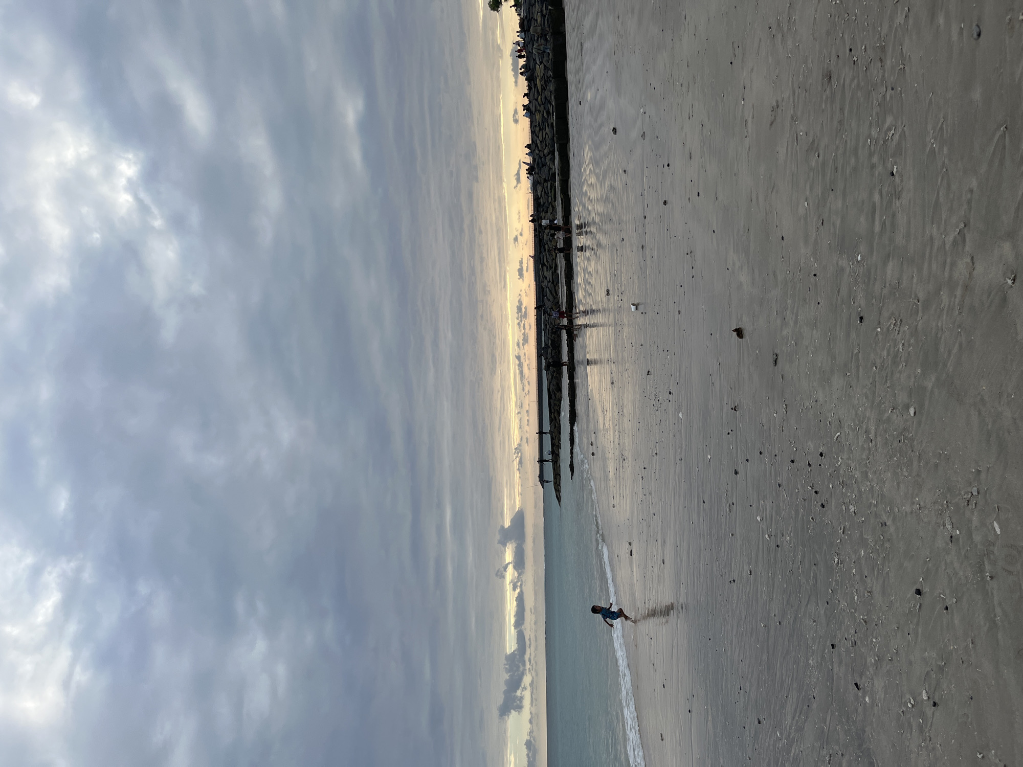 A lone child runs on the wet sand near the ocean's edge. The sky is overcast with heavy clouds, but a faint glow of sunset is visible on the horizon. People are gathered on a rocky pier in the distance, some appearing to be fishing. The beach is a mix of wet and dry sand, with small shells and debris scattered across the surface. The scene is calm and serene, with a sense of quiet activity.