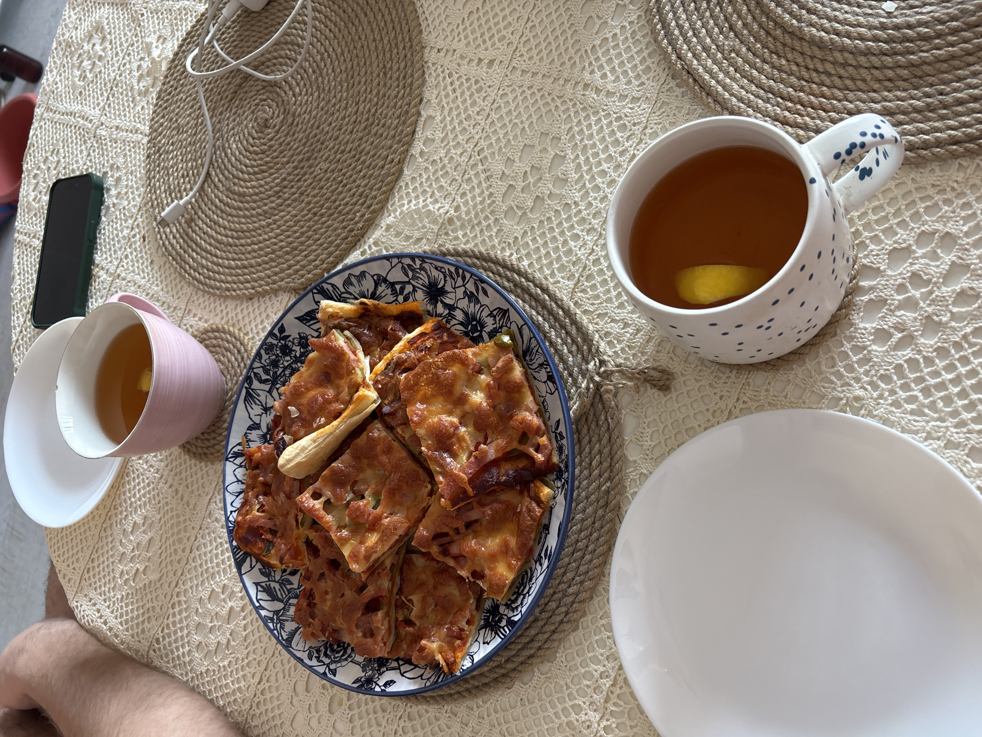 The scene depicts a casual meal setting on a tablecloth with a knitted pattern, likely indoors. The location cannot be identified as Krasnoyarsk, Russia, based solely on visual information. A plate of square-cut pizza pieces is the central element. To the left is a pink teacup and saucer filled with tea and a lemon slice, along with a smartphone and a charging cable. To the right is a larger white teacup with tea and a lemon slice and a plain white plate. Each teacup sits on a round, braided placemat. A leg, presumably belonging to a person, is visible at the bottom left of the frame.