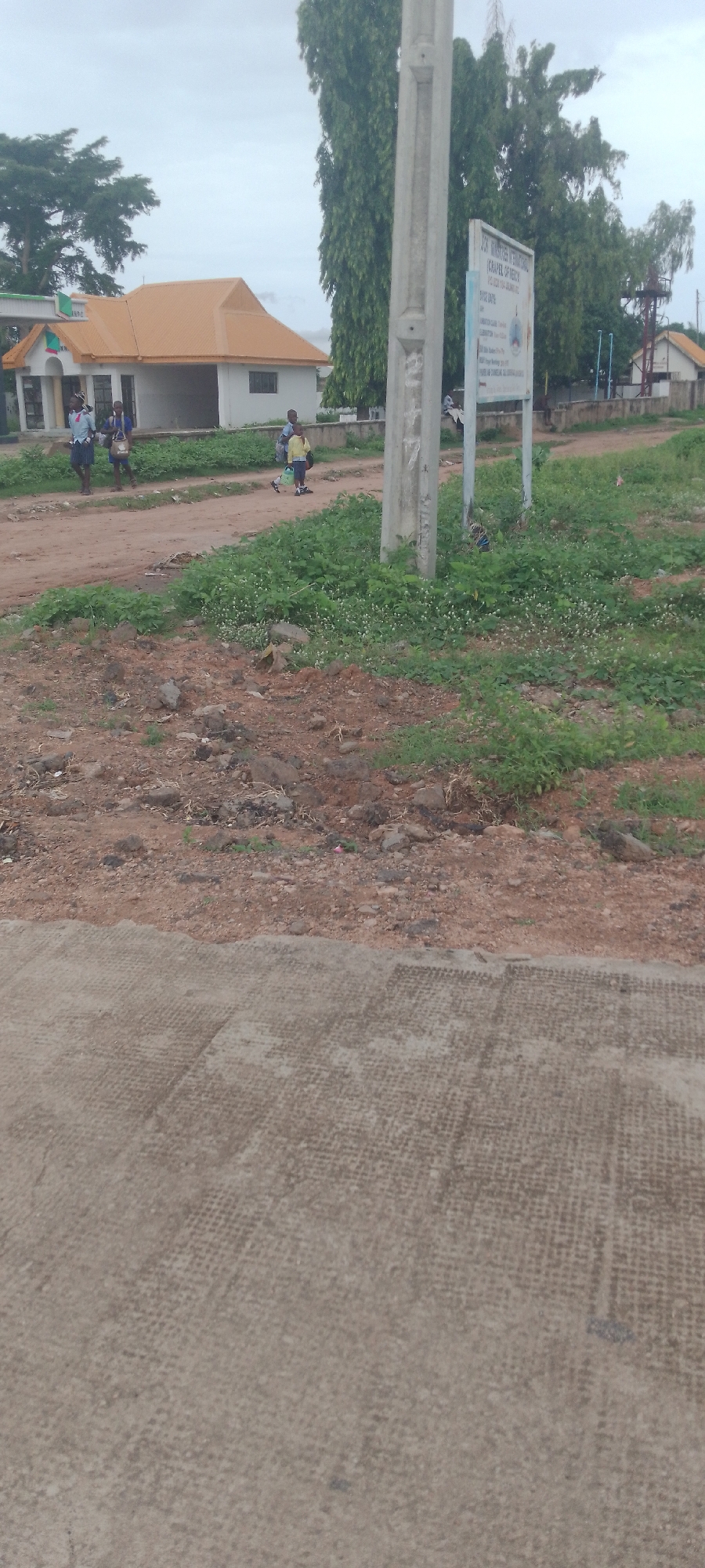 The image depicts a cloudy day in Jalingo, Nigeria, likely near a school or religious institution, evidenced by a sign that reads "JSS JUBILEE INTERNATIONAL (CHAPEL OF MERCY) P.O. BOX 154 JALINGO NG". A single concrete lamppost stands prominently in the foreground, to the right of center. Behind it, a group of children, possibly students, are seen walking. One child in the front left wears a light blue shirt and dark skirt, carrying a bag. Another child next to them wears a dark blue shirt and also carries a bag. Further back, to the right, a child in a yellow shirt and dark shorts walks with a light-colored bag.
The background features a light-colored building with an orange-tiled roof, suggestive of a modern structure. To the far right, another smaller building with a similar roofline is visible, along with what appears to be a water tower. A fence runs along the property line. The ground is a mix of dirt, gravel, and sparse green vegetation with small white flowers. The foreground shows a concrete surface with a textured pattern, possibly a sidewalk or pavement.