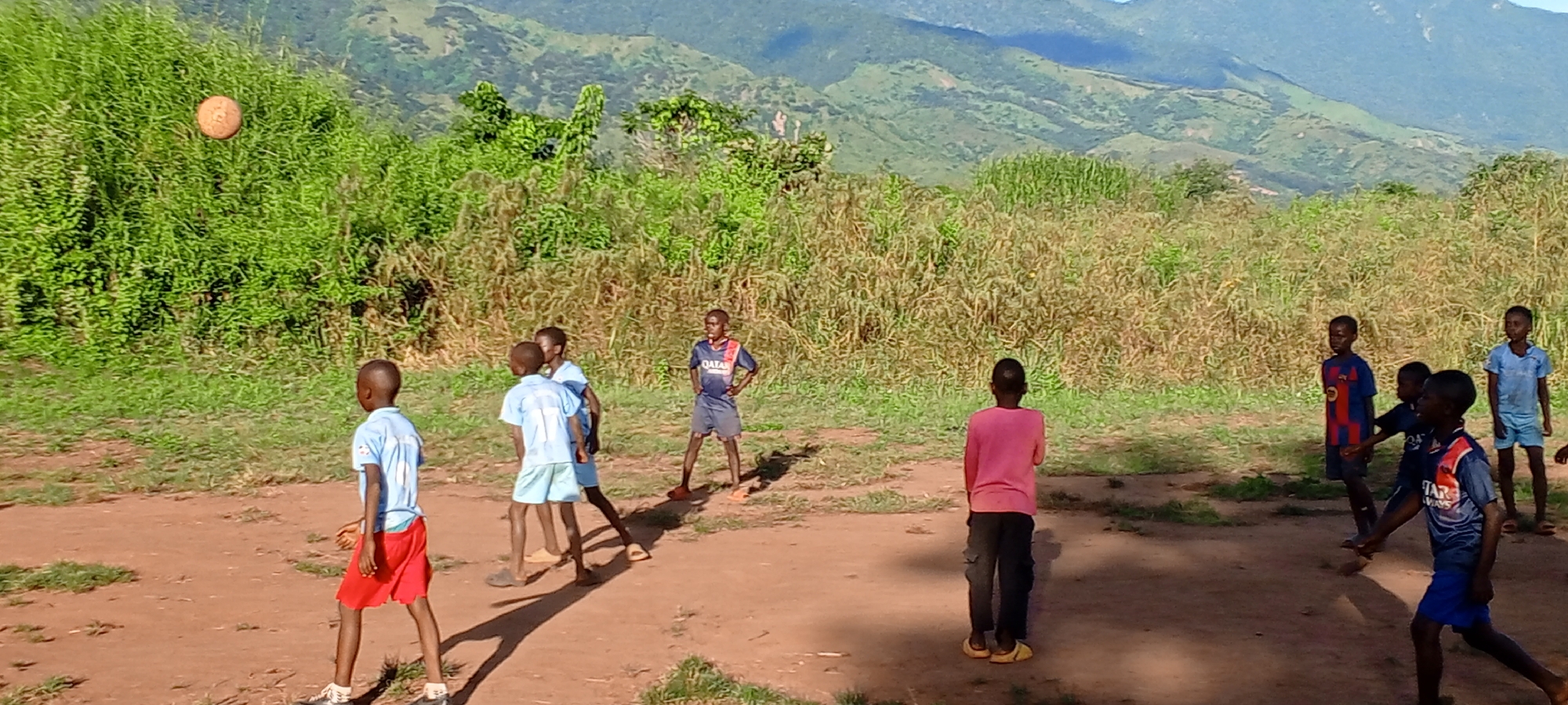 In Ngalula, Democratic Republic of the Congo, a group of children play on a dirt field with lush greenery and distant mountains in the background. A soccer ball is in mid-air near the vegetation on the left. Among the children, some are wearing soccer jerseys, while others wear casual clothing, indicating they are engaged in an informal game. They appear to be interacting, possibly playing soccer. The scene conveys an ordinary moment of children's recreation in a rural or semi-rural setting.