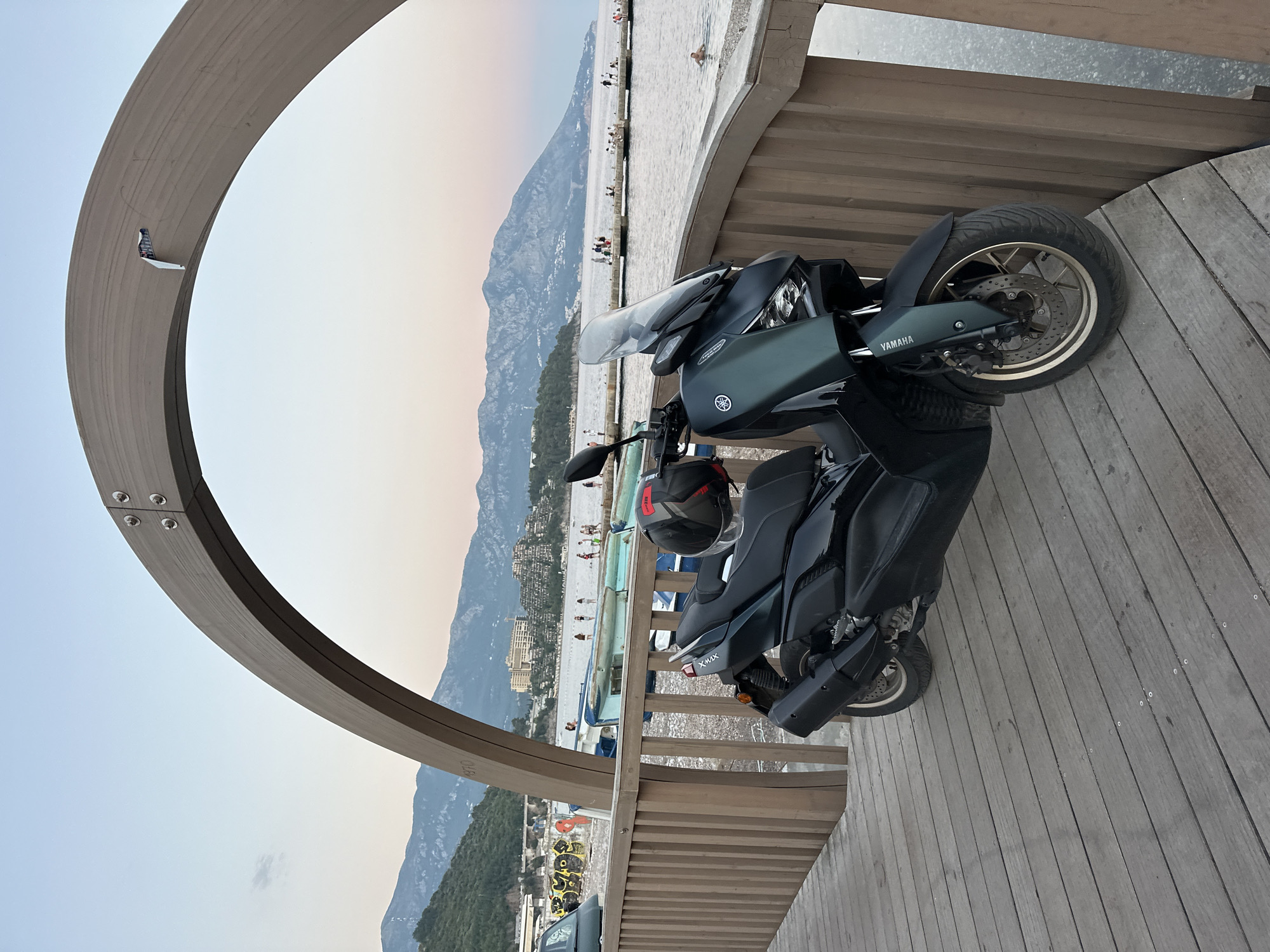 A dark teal Yamaha XMAX scooter is parked on a wooden boardwalk. The boardwalk is part of a structure that includes a large wooden arch overhead. In the background, there is a calm body of water, a beach with people, and a town built into a hillside. Mountains are visible in the distance, and the sky is a gradient of pale blue and pink, indicating it is either dawn or dusk. The scooter is positioned to the right of the frame, with its front wheel angled slightly towards the viewer. A black helmet with red accents rests on the seat of the scooter. The wooden planks of the boardwalk are weathered and run diagonally across the foreground. The town in the background features buildings with red-tiled roofs and a few larger, modern hotel-like structures.