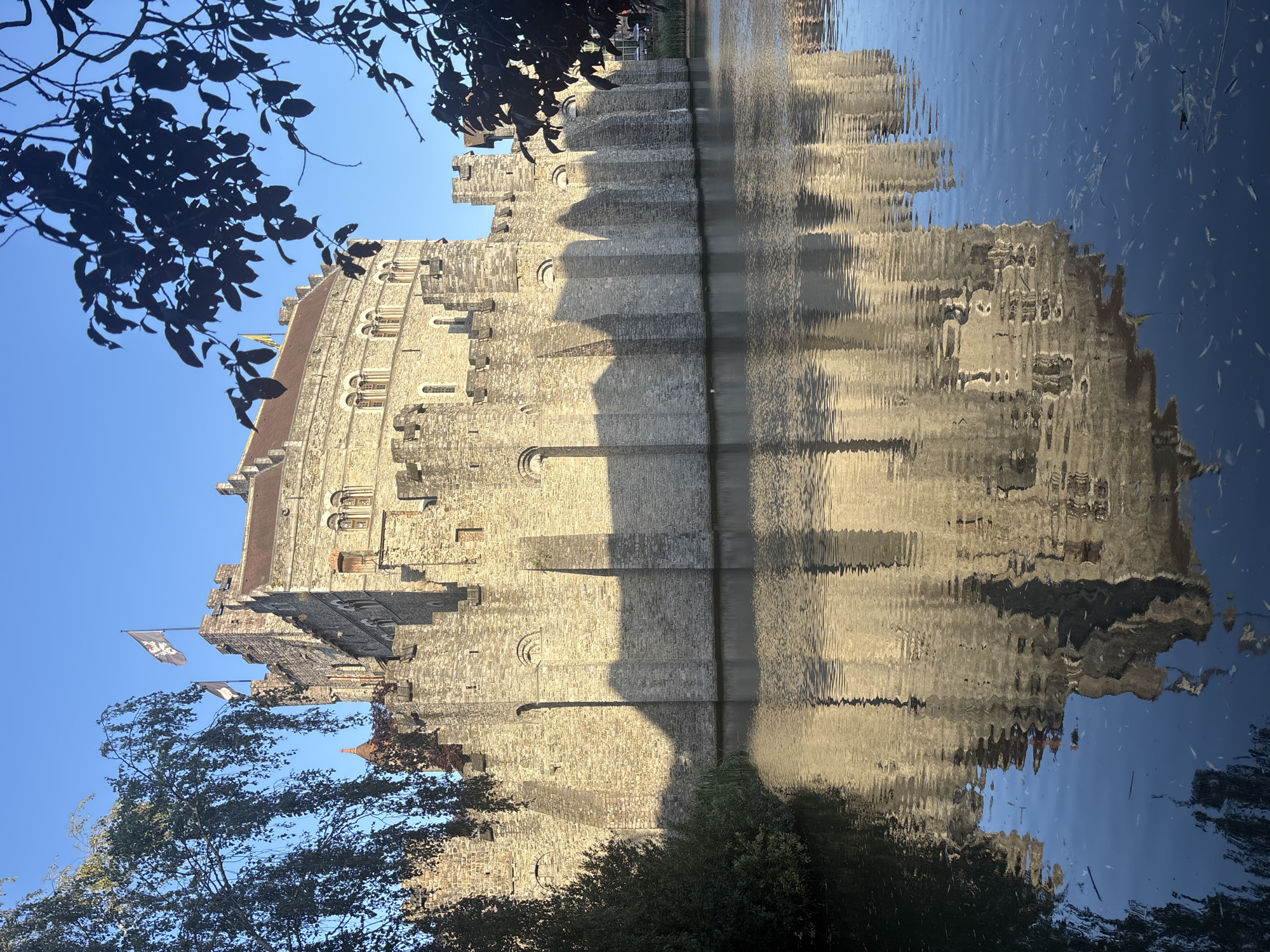The image shows the Gravensteen Castle in Ghent, Belgium, on a clear day. The stone castle, with its crenellated battlements and turrets, is surrounded by a moat. The sun casts long shadows on the castle walls, and the clear blue sky provides a vibrant backdrop. The reflection of the castle is visible in the calm water of the moat. Trees and foliage frame the right and left sides of the image, adding a touch of nature to the historic scene. No people are visible in the image.