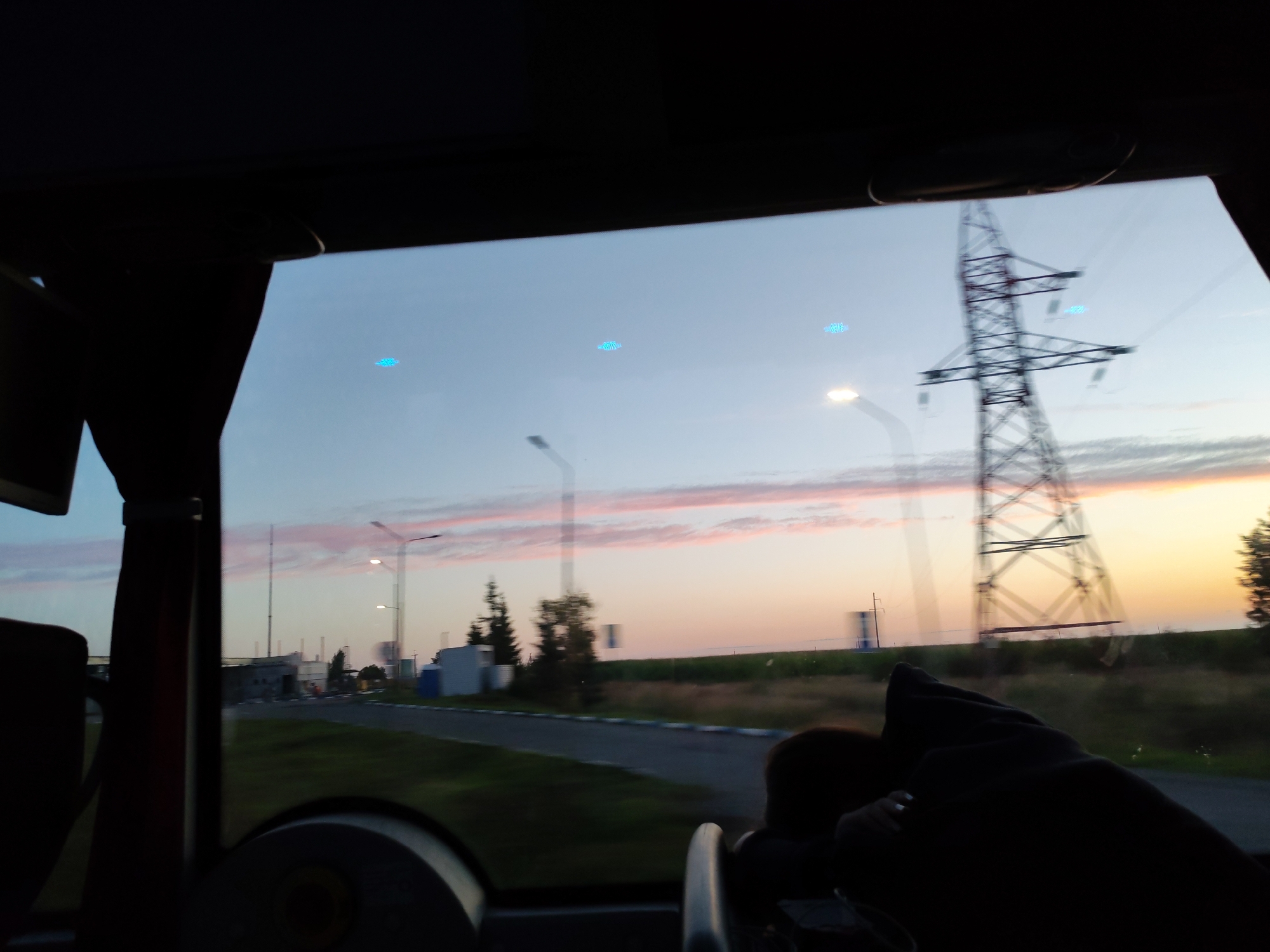 The image captures a view through a bus window at dusk. In the foreground, the dark interior of the bus is visible, with parts of seats and a person's arm resting on a dark fabric. The middle ground shows a road with a sidewalk, flanked by grass and some industrial-looking buildings and trees. In the background, a large electrical tower dominates the scene, with a pale sky transitioning from light blue to a soft orange and pink as the sun sets. There are a few indistinct glowing blue objects in the sky, possibly reflections or lens flares. The overall impression is a somewhat blurred, moving scene of an industrial area at twilight.