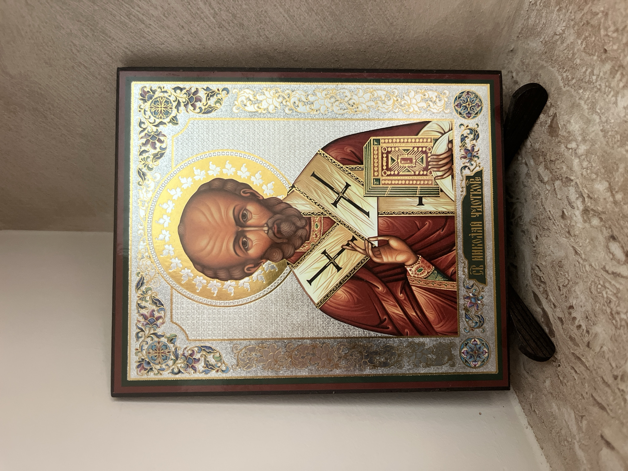 The image shows a rectangular religious icon in a brown frame. The icon depicts a figure with a bald head and beard, wearing a red and gold robe with a cross. A golden halo surrounds his head. He holds a book in his left hand and is gesturing with his right. The background is silver with ornate floral designs in the corners. Cyrillic text is visible on the right side of the icon. The icon is mounted on a wall where the surfaces vary: one side appears to be a smooth off-white, while the other is a textured, beige stone. A dark, narrow object is positioned between the icon and the textured wall. The scene is likely in a church or religious setting in Varna, Bulgaria.