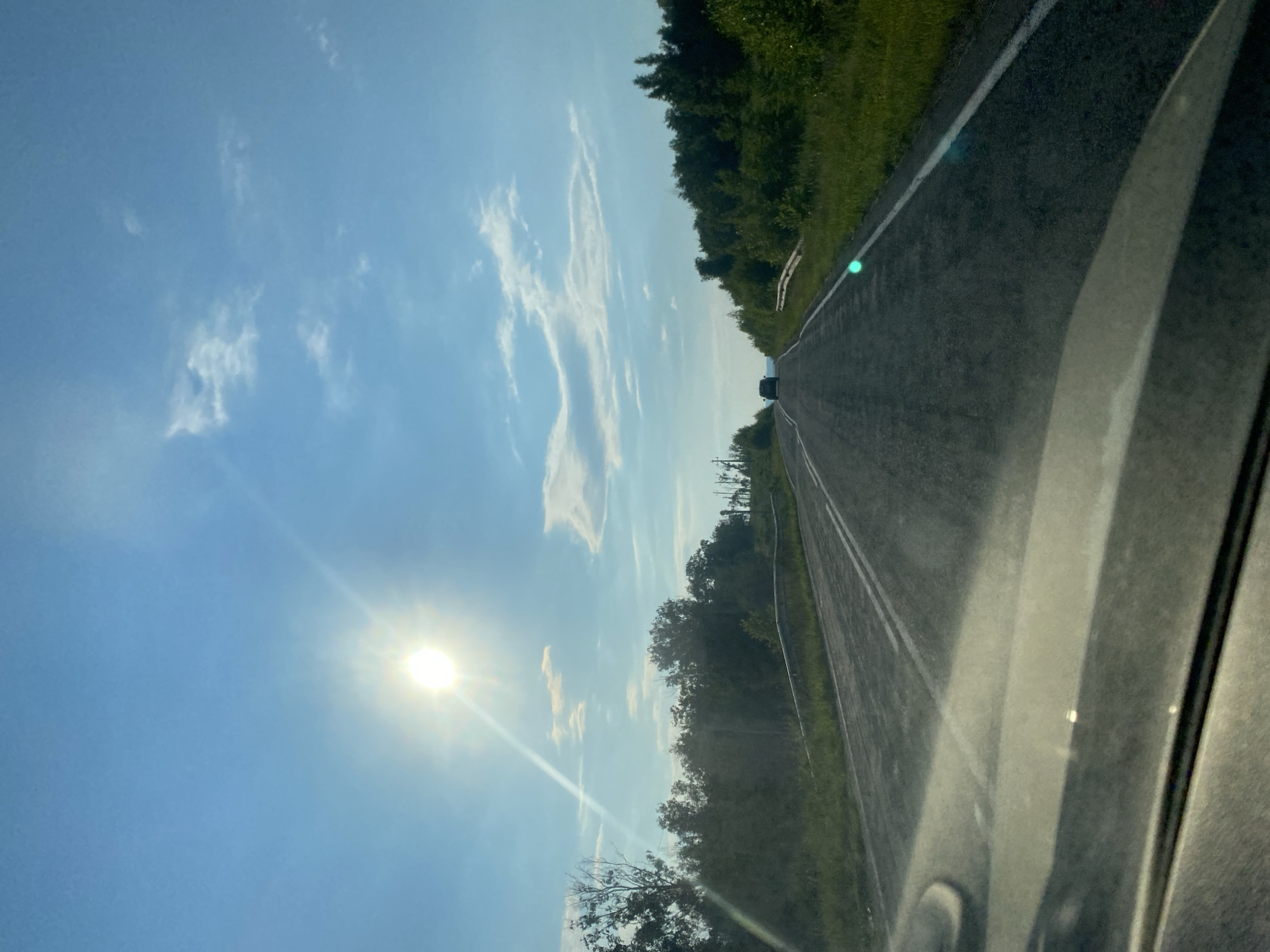 The image depicts a view from inside a vehicle traveling on a paved road, likely in the city of Antingan, Russia. The sun is prominently visible in the upper left quadrant of the sky, casting a bright glare and creating lens flare effects across the image. The sky is a clear blue with scattered white clouds. The road is asphalt, with white lane markings visible. On either side of the road, there is dense green vegetation, consisting of trees and bushes. A guardrail runs along the right side of the road. In the distance, a dark-colored car is visible on the road. The foreground shows the dashboard and windshield of the vehicle, with reflections and glare from the sun. The overall scene suggests a sunny daytime drive through a rural or semi-rural area.