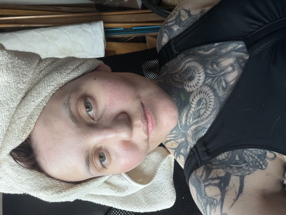 A person with fair skin and light eyes faces the camera, a white towel wrapped around their head. They wear a black sports bra, revealing intricate snake tattoos covering their chest, neck, and shoulder. The background includes a blurred wooden surface and a white rectangular object, possibly a cushion or towel. The image is well-lit and the subject appears relaxed.
