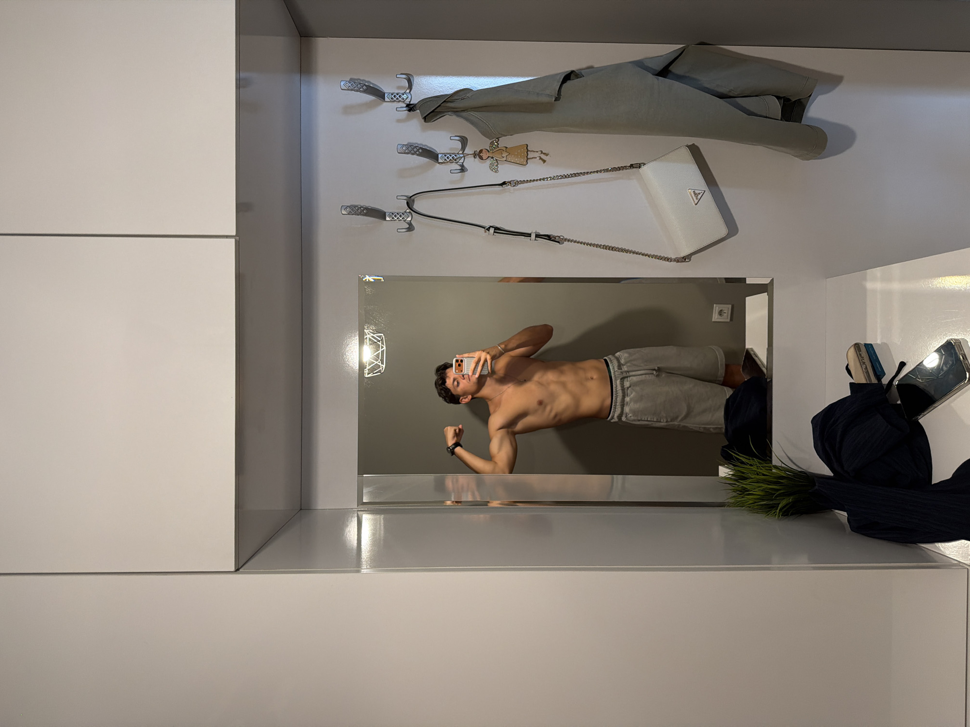 A young adult male is taking a mirror selfie in what appears to be a modern hallway or entryway. He is shirtless, wearing gray shorts, and flexing his bicep. He is holding a smartphone in his right hand to capture the photo. To his right, on the wall, are several coat hooks. A white handbag with a silver chain strap is hanging from one of the hooks, along with a light green jacket or sweater. A small, whimsical angel charm is also attached to one of the hooks. Below the mirror, on a white surface, a dark blue garment, possibly a sweater or scarf, is visible along with a plant in a dark pot. A smartphone is also resting on this surface. The environment features clean, minimalist lines with white cabinetry. No specific landmarks of Blagoevgrad, Bulgaria, are identifiable within this indoor setting.