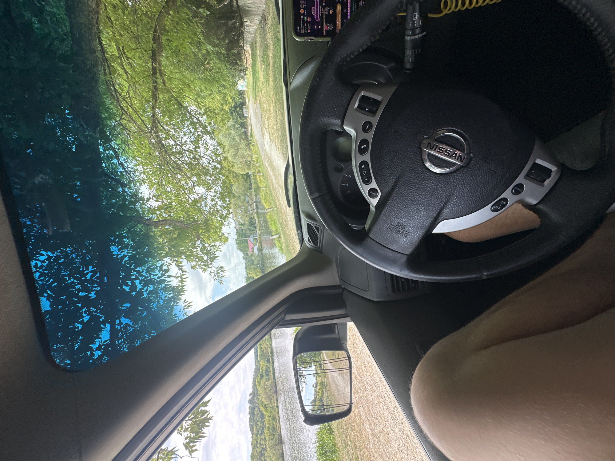 The image depicts the interior of a car, viewed from the driver's perspective. The driver's left leg and knee are visible in the lower left portion of the frame. The steering wheel, with a Nissan logo, is centered in the foreground. The car's dashboard and a phone mounted on the right side are also visible. Outside the car, through the windshield and side window, a body of water, a dirt road, and lush green trees are seen. The scene appears to be in Makiv, Ukraine, given the natural landscape of trees and water. The time of day appears to be daytime, with sunlight filtering through the leaves.