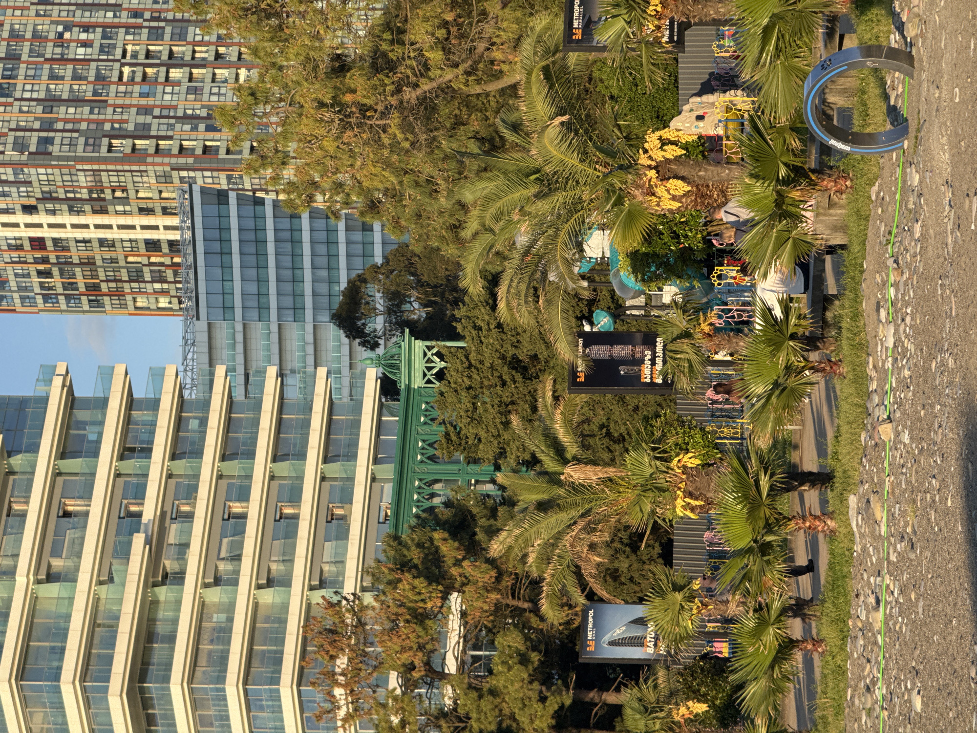 The image depicts an urban scene, likely a park or promenade in Batumi, Georgia. Buildings of modern architecture dominate the background. To the left is a mid-rise structure with a facade of glass and vertical columns. To the top is a tall, multi-storied building. In the foreground are lush trees and palm trees. A playground is visible amidst the greenery. A metal sculpture stands on the edge of a paved path. Park infrastructure, such as signs, is dispersed throughout the scene. Two people can be seen in the playground. The environment is sunlit, indicating it is likely daytime.