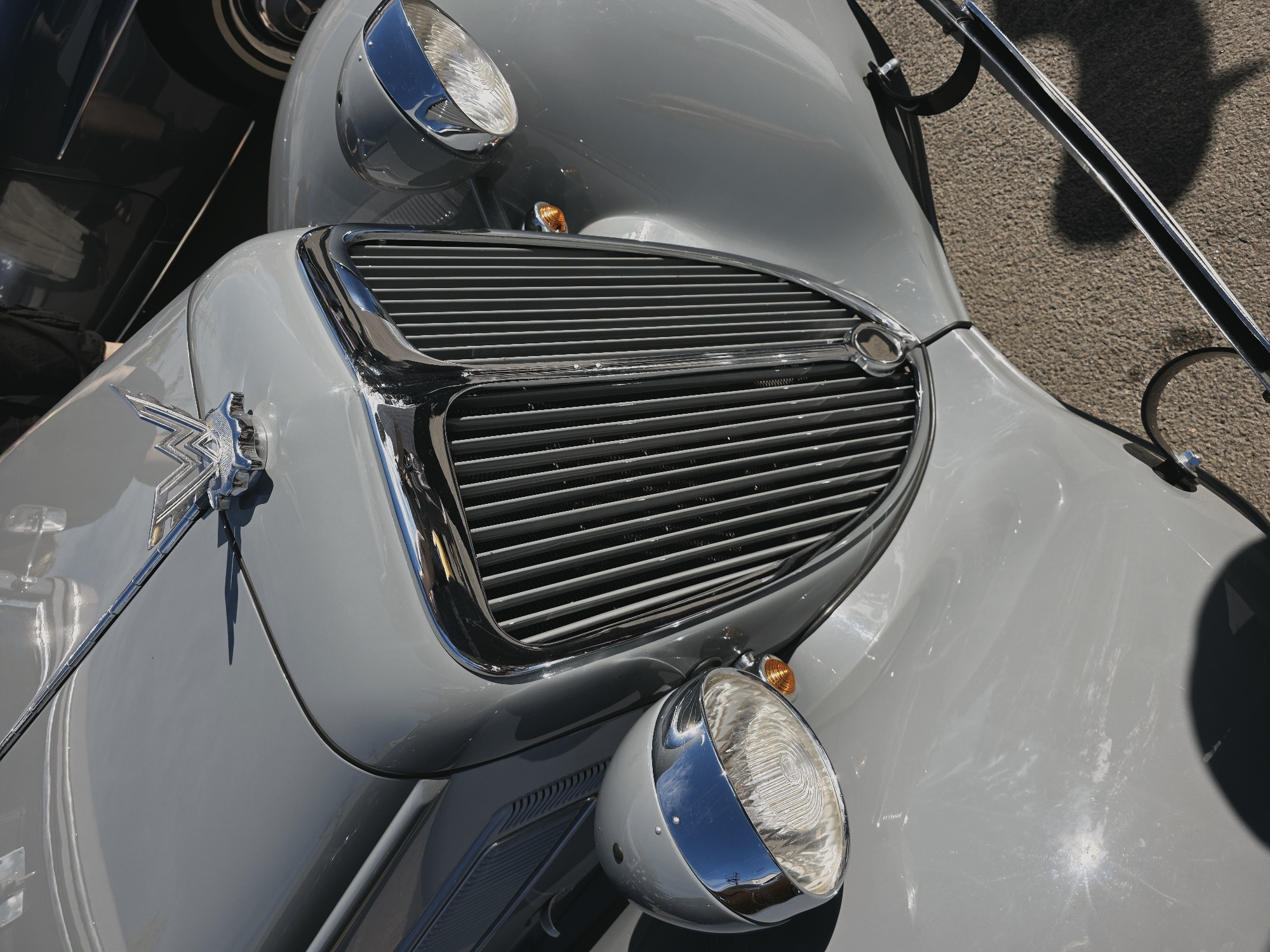 This is a close-up, eye-level, outdoor, daylight, vertical, full shot of the front of a vintage, light grey automobile. The car's grille is prominent, featuring vertical chrome-plated bars. Two round, chrome-accented headlights with glass lenses are visible on either side of the grille, along with small amber turn signals. A chrome hood ornament, shaped like a stylized "W" with wings, is centered on the hood above the grille. The car's paint shows some scuffs and scratches. The background reveals a dark-colored vehicle and a paved surface, possibly asphalt or gravel. The scene is likely located in Saint Petersburg, Russia, given the context of similar images, but there are no explicit visual cues within this image to confirm the location. No people are visible.