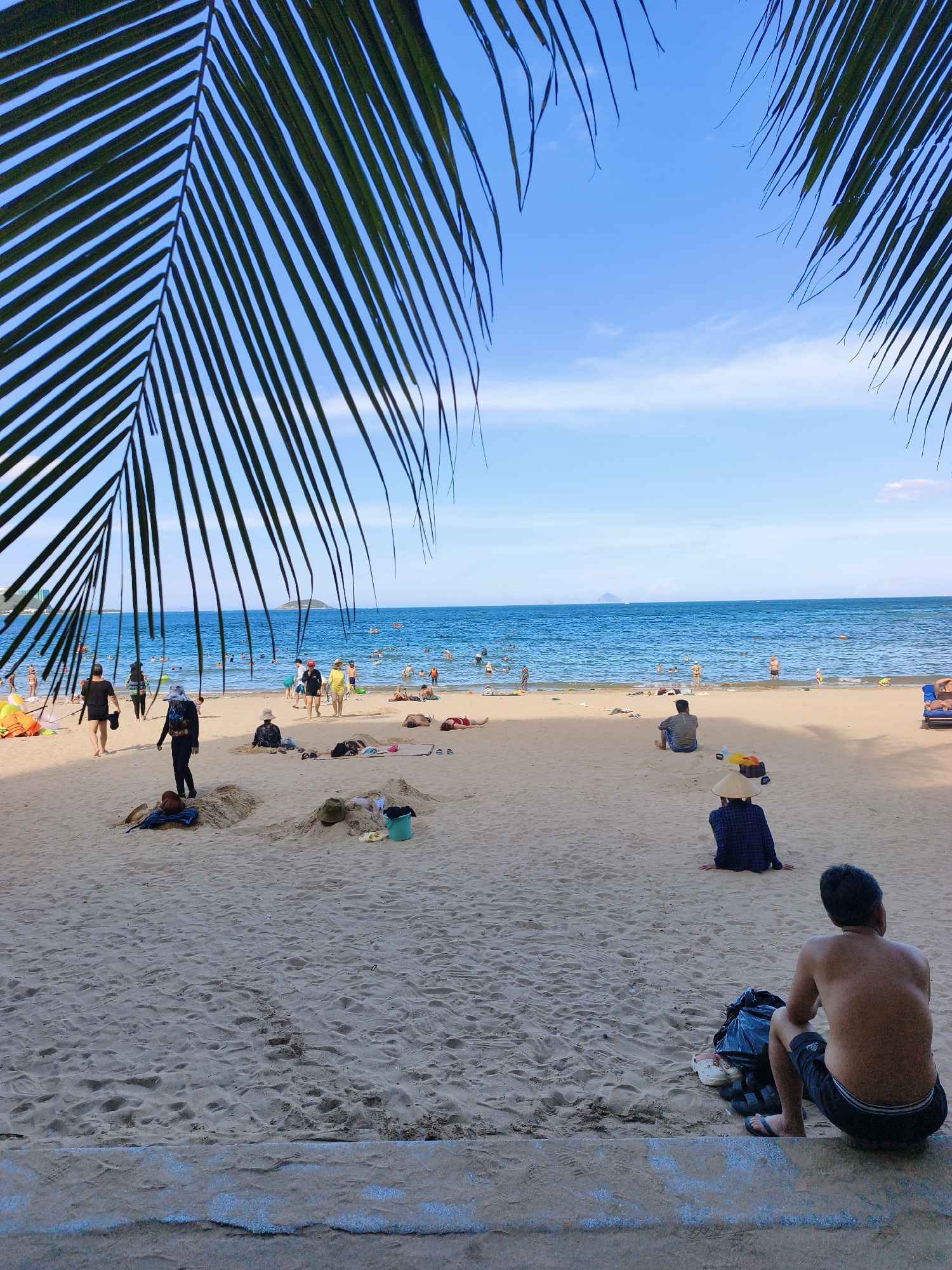This image depicts a crowded beach in Nha Trang, Vietnam, likely located along the central coastline. In the foreground, a person with their back to the camera sits on a concrete ledge, their torso bare, wearing black shorts and flip-flops. Next to them is a black trash bag and a pair of shoes. A conical Vietnamese hat rests on the sand nearby. Further up the beach, several people are seated in the sand, some sunbathing, while others are dressed in casual beach attire. A few individuals are walking towards the water, which is filled with swimmers and people wading. The beach itself is sandy, with small mounds of sand scattered across it. Palm fronds frame the top of the image, casting shadows and suggesting a tropical setting. The ocean is a vibrant blue, with a clear sky overhead. The overall scene conveys a lively and typical day at a popular beach resort.