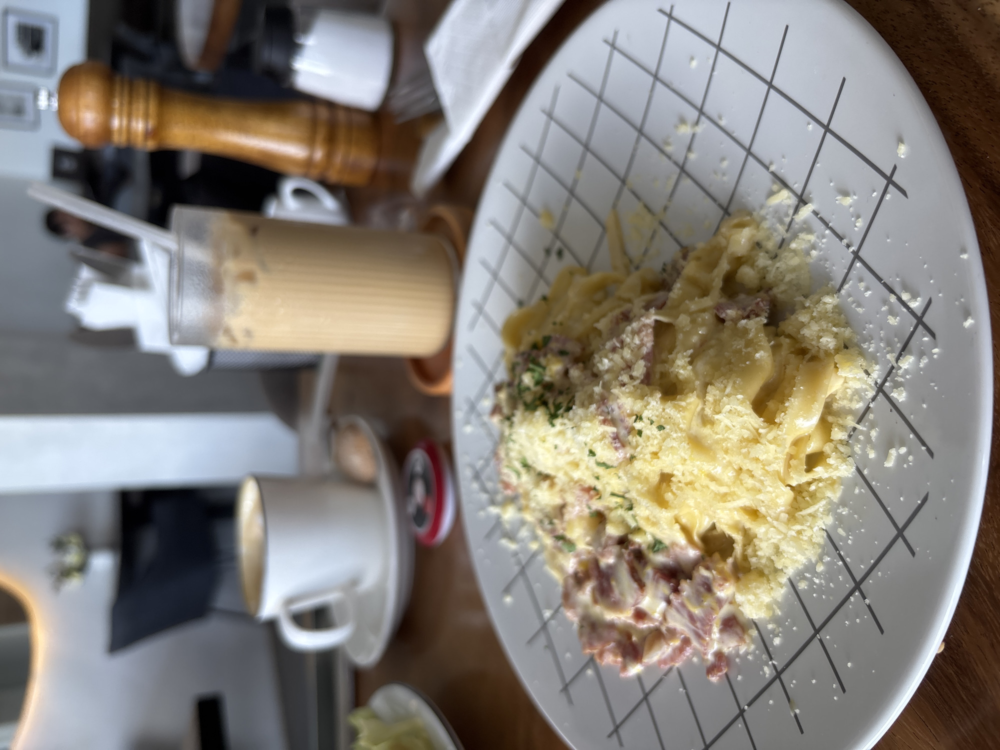 The scene depicts a table setting in Kuta Selatan, Indonesia. A plate of pasta, seemingly carbonara, dominates the view, liberally sprinkled with grated cheese. To the left, a tall glass of iced coffee sits near a white ceramic mug, implying a beverage service. A wooden pepper grinder is positioned above the coffee. The plate's design is a grid of black lines. The surrounding environment is blurred, suggesting an indoor setting in a cafe or restaurant.