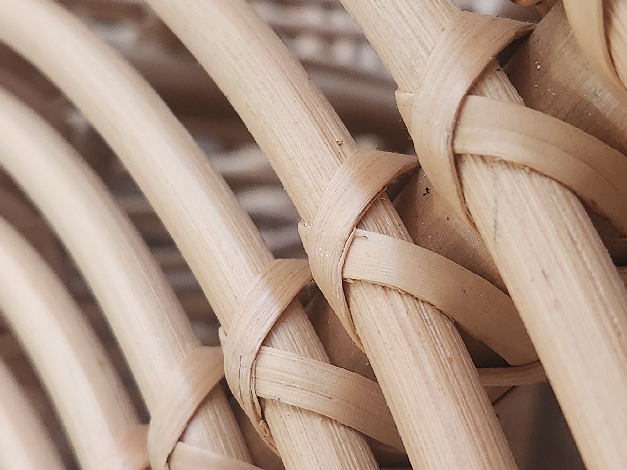 This is a close-up, high-angle shot of woven natural material, likely rattan or bamboo, forming a textured pattern. The strands are light brown and cylindrical, interlaced with flatter, wider strands that create a diamond-like weave in places. The lighting is bright, highlighting the natural grain and subtle variations in color of the material. The background is blurred, suggesting it is either further sections of the same woven material or a neutral, out-of-focus surface. There is no discernible activity or event taking place; the image focuses solely on the texture and structure of the woven object. No people, text, or specific location cues are visible, making it difficult to determine the exact setting beyond the material itself. The overall impression is one of natural texture and handcrafted detail.