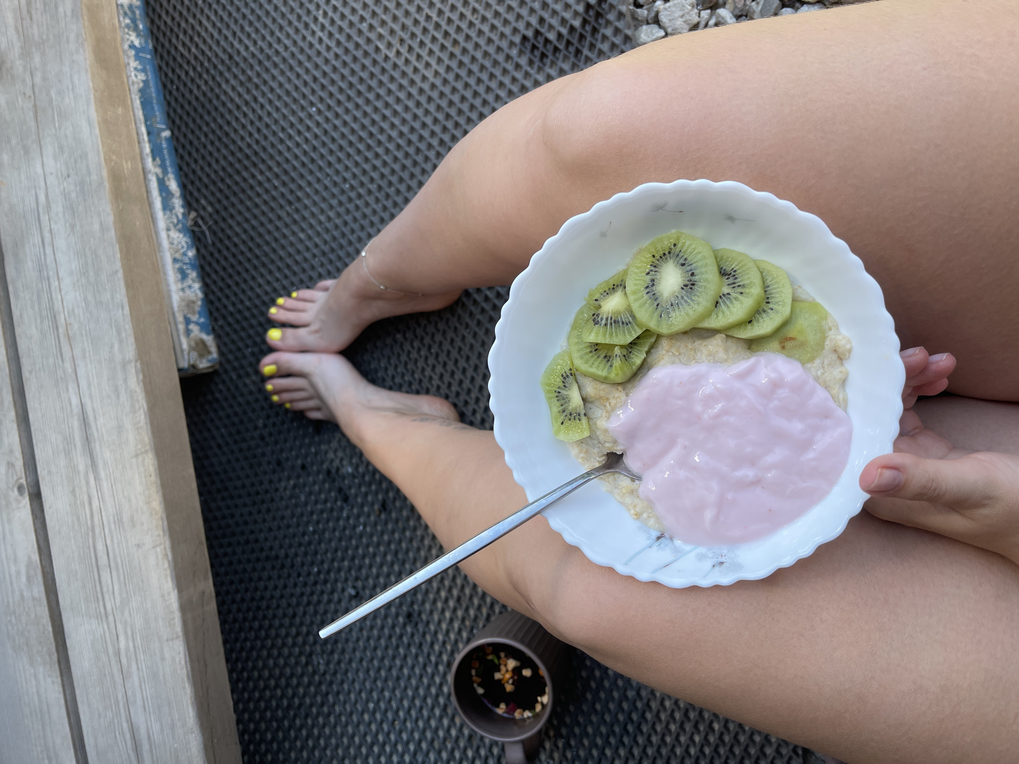 A person is seated outdoors with a bowl of oatmeal topped with yogurt and kiwi slices. A cup of coffee or tea sits to the left of the bowl. The person's bare feet, painted with yellow nail polish, are visible in the foreground. The background consists of a wooden bench and a dark, textured surface. The image does not provide any information about the city of Мосметаллоконструкция Онт, Russia.