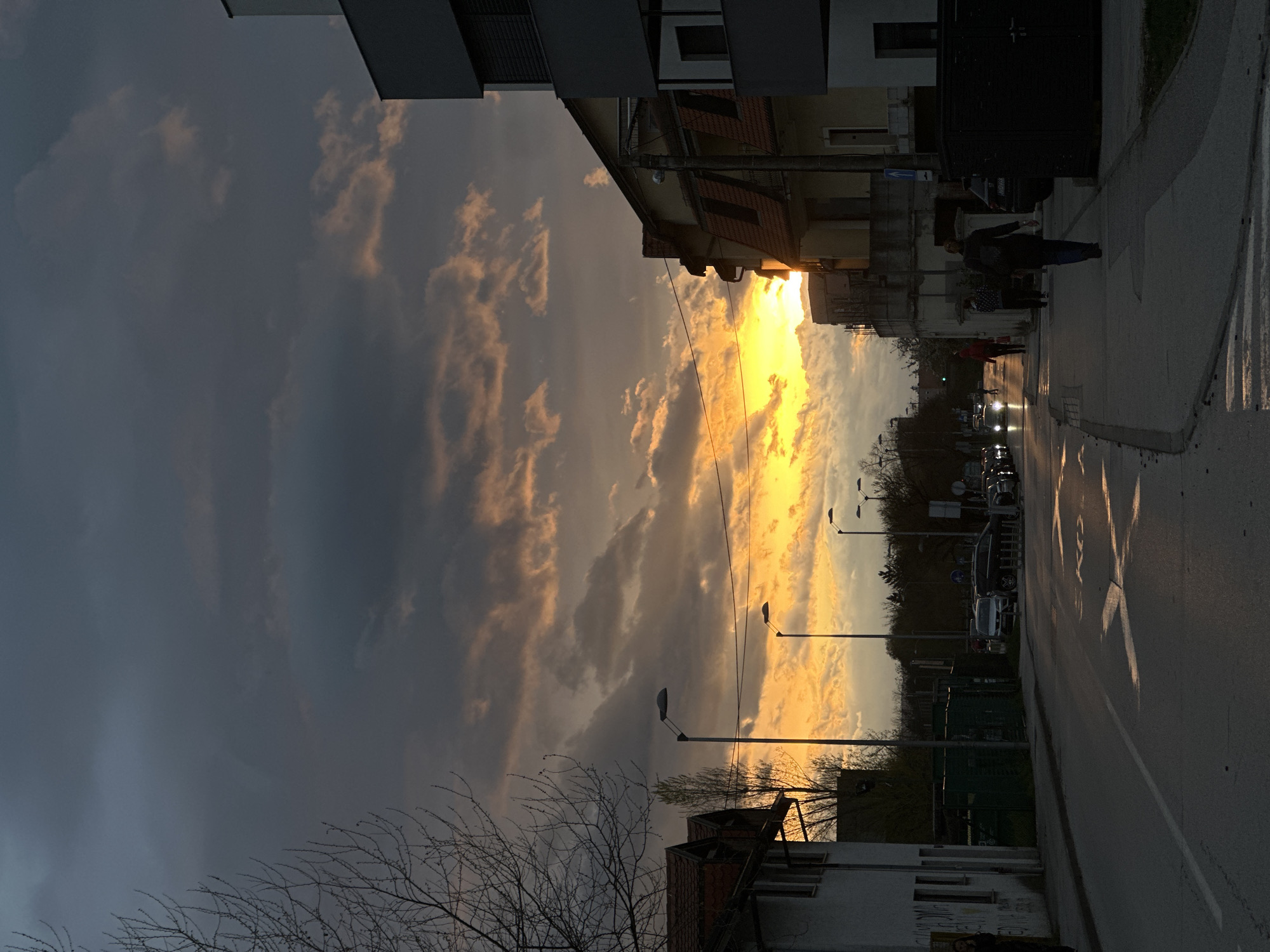 The image depicts an urban street scene during sunset in the City of Zagreb, Croatia. The sky is divided between dark storm clouds and brilliant orange and yellow hues emanating from the setting sun. The sun appears to be setting down the street, between buildings.
On the right side of the image is a sidewalk with a zebra crossing painted on the street. People are visible walking on the sidewalk. Cars are parked and driving down the street. Streetlights are lined along the road. Buildings of varying heights are on either side of the street. A bare tree is in the lower left corner.