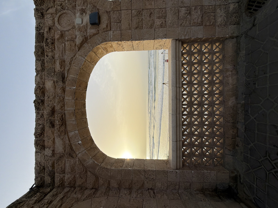 The image displays a picturesque scene framed by an architectural opening made of rough-hewn, light-colored stone blocks, likely depicting a historical or rustic structure. The opening consists of a semi-circular arch on the left and a more angular, rectangular section on the right, which is partially filled with a decorative lattice screen.
Through this opening, a vibrant view of a beach and the ocean is visible during either sunrise or sunset. The sun is low on the horizon, partially obscured by the bottom edge of the arch, casting a bright, warm yellow and orange glow across the sky and reflecting on the water. The sky transitions to a lighter, softer blue-grey higher up.
The ocean extends horizontally across the frame, with gentle waves breaking on the sandy shore. On the beach, small figures appear as silhouettes against the bright sky, indicating human presence. There seem to be at least two figures, one closer to the water's edge and another further along the shore. One figure near the water appears to be holding a long, thin object, possibly a fishing rod or pole. A small red object, possibly a flag or marker, is also visible on the beach to the right of the figures.
The stone structure itself shows intricate details. The blocks are varied in size and shape, creating a textured surface. On the upper left part of the wall, above the arch, there's a circular relief carving and a rectangular light fixture or box mounted to the wall. The right side of the opening features a decorative screen made of square concrete or stone blocks, each containing an "X" or cross-shaped cutout, forming a repetitive geometric pattern. Sunlight filters through these cutouts, highlighting their form. The ground or floor area visible beneath this screen and to the far right is paved with dark, patterned tiles.
The overall impression is one of tranquility and beauty, capturing a peaceful moment by the sea viewed from within an ancient-looking edifice.