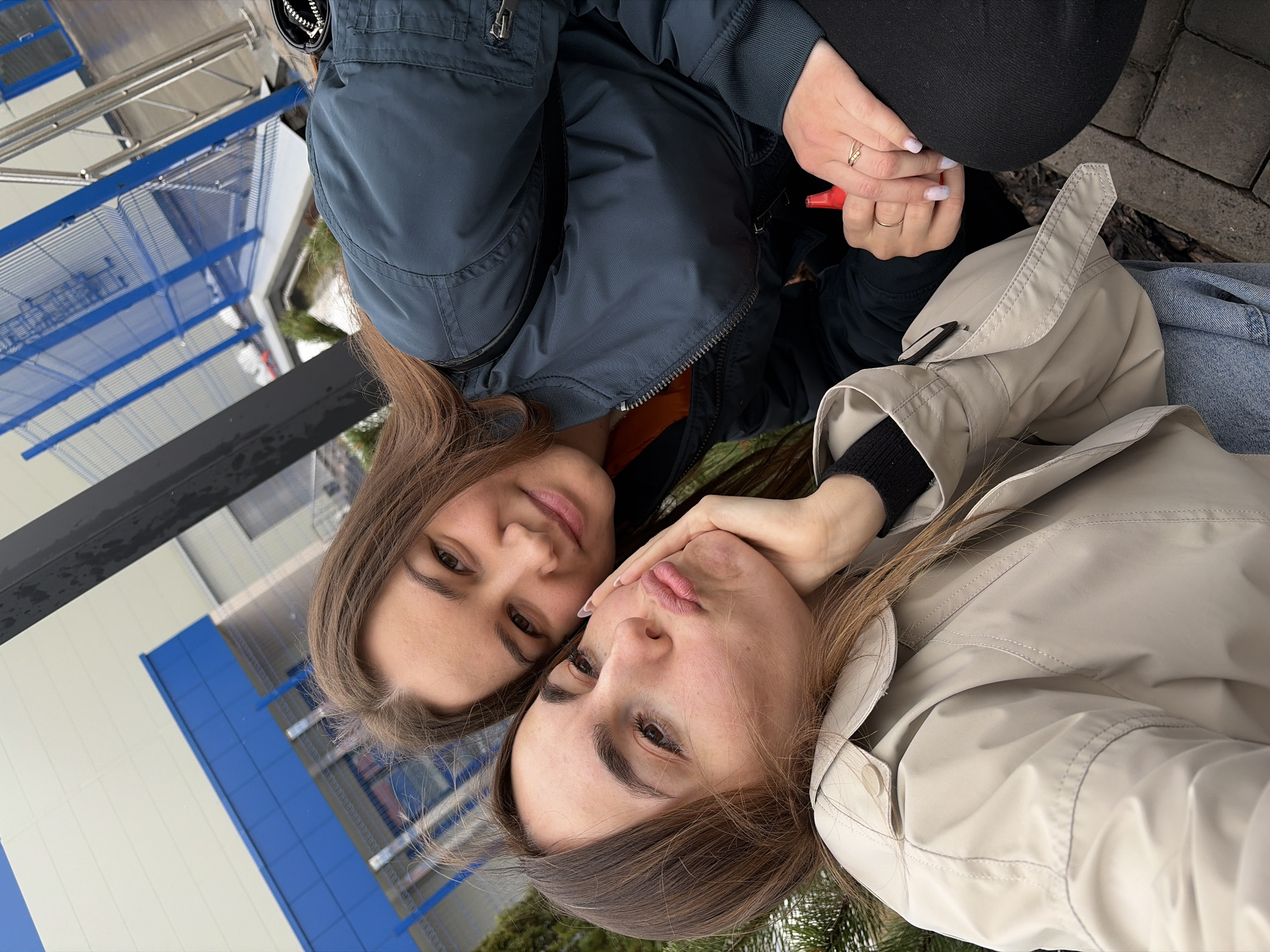 Two women are closely positioned, likely taking a selfie. One woman is wearing a dark blue puffer jacket with a visible orange underlayer and black bottoms. She is holding a red object in her hand. The other woman is wearing a beige trench coat over a black sweater. The location appears to be urban, possibly in Izhevsk, Russia, judging by architectural elements such as the blue-paneled building and metal scaffolding in the background. The ground is paved with rectangular stones.