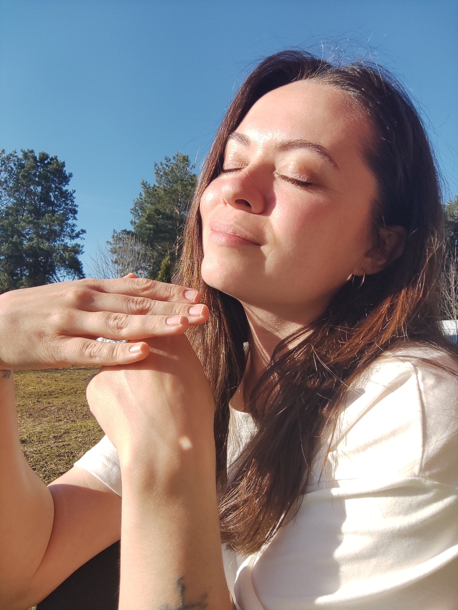 A serene portrait captures a woman with closed eyes, basking in bright sunlight in an outdoor setting. The woman, appearing to be in her 20s or 30s, has long dark brown hair and a peaceful expression, with her face gently tilted upwards towards the sun. Her skin is warmly illuminated by direct sunlight, creating highlights on her forehead, nose, and cheeks, and casting soft shadows that emphasize the strong light source.
She is wearing a light-colored, likely white or off-white, t-shirt. Her hands are clasped together in front of her, resting comfortably on her arm, revealing neat, unpolished fingernails and a ring on one finger. Small hoop earrings are visible in her ears. A faint tattoo is partially visible on her left wrist.
The background features a clear, vibrant blue sky with no clouds. Several tall evergreen trees, appearing to be pines, are visible, along with some bare branches from deciduous trees in the distance, suggesting early spring or late autumn. The ground is covered in green and brown grass or foliage, indicating a natural environment such as a park or countryside. The strong sunlight and clear sky point to a bright, sunny day, likely mid-morning or afternoon. The overall mood conveyed is one of tranquility, relaxation, and a moment of enjoying nature and the sun's warmth. No visible text is present in the image. The location context for this scene is Фоки, Russia.