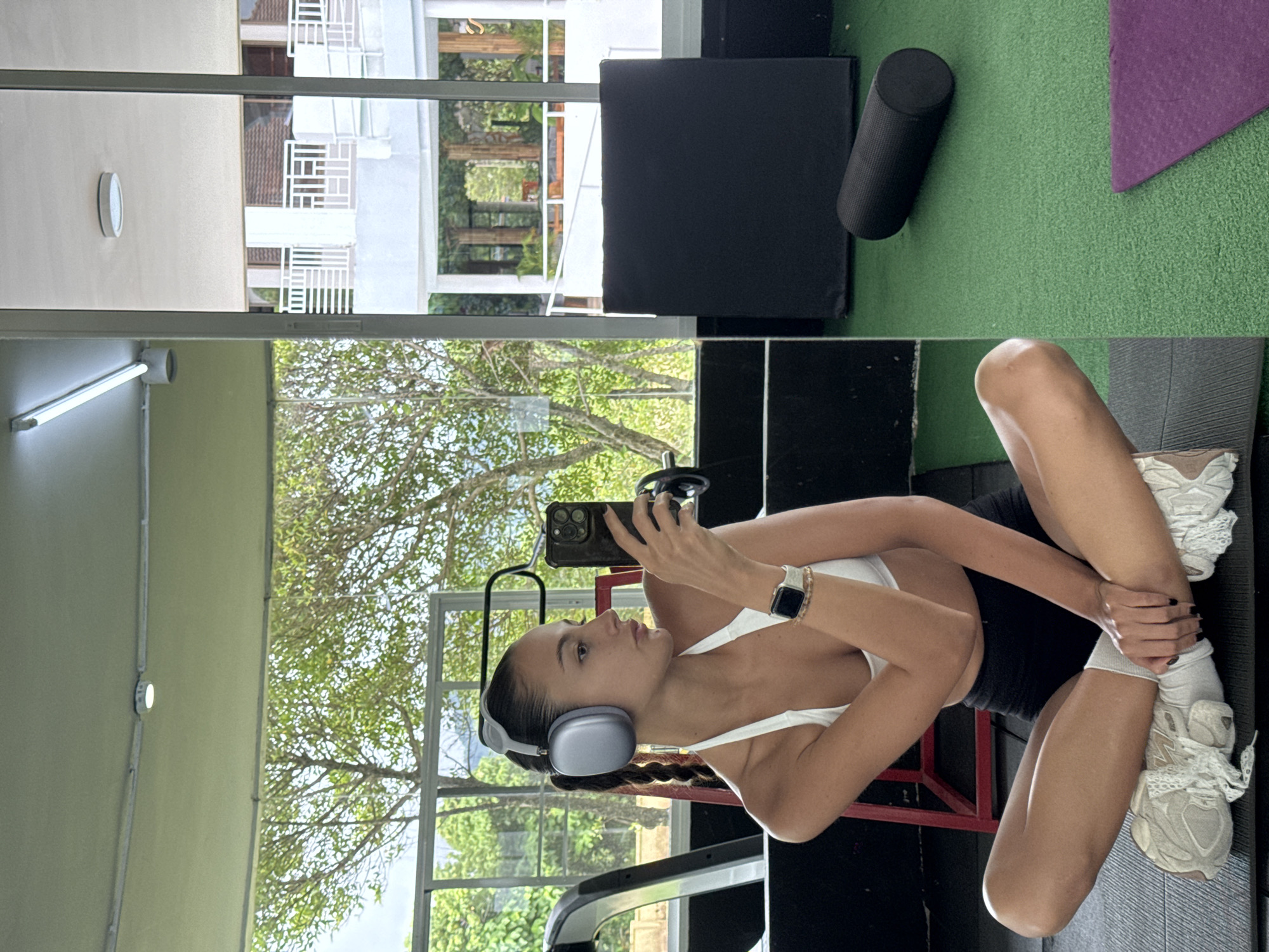 The image captures a young woman indoors, likely within a fitness or recreational facility. She is seated with crossed legs, holding a smartphone and apparently taking a photo or video of herself. She wears a white sports bra, black shorts, white socks, and running shoes. Additionally, she has headphones and a wristwatch. A large window in the background reveals a lush green landscape, consistent with tropical vegetation. Further details visible through the window suggest a building complex, possibly residential or commercial. Given the information, this image was taken in the city of Kathu, Thailand. Fitness equipment and gym accessories are also present in the background, corroborating the gym environment.
