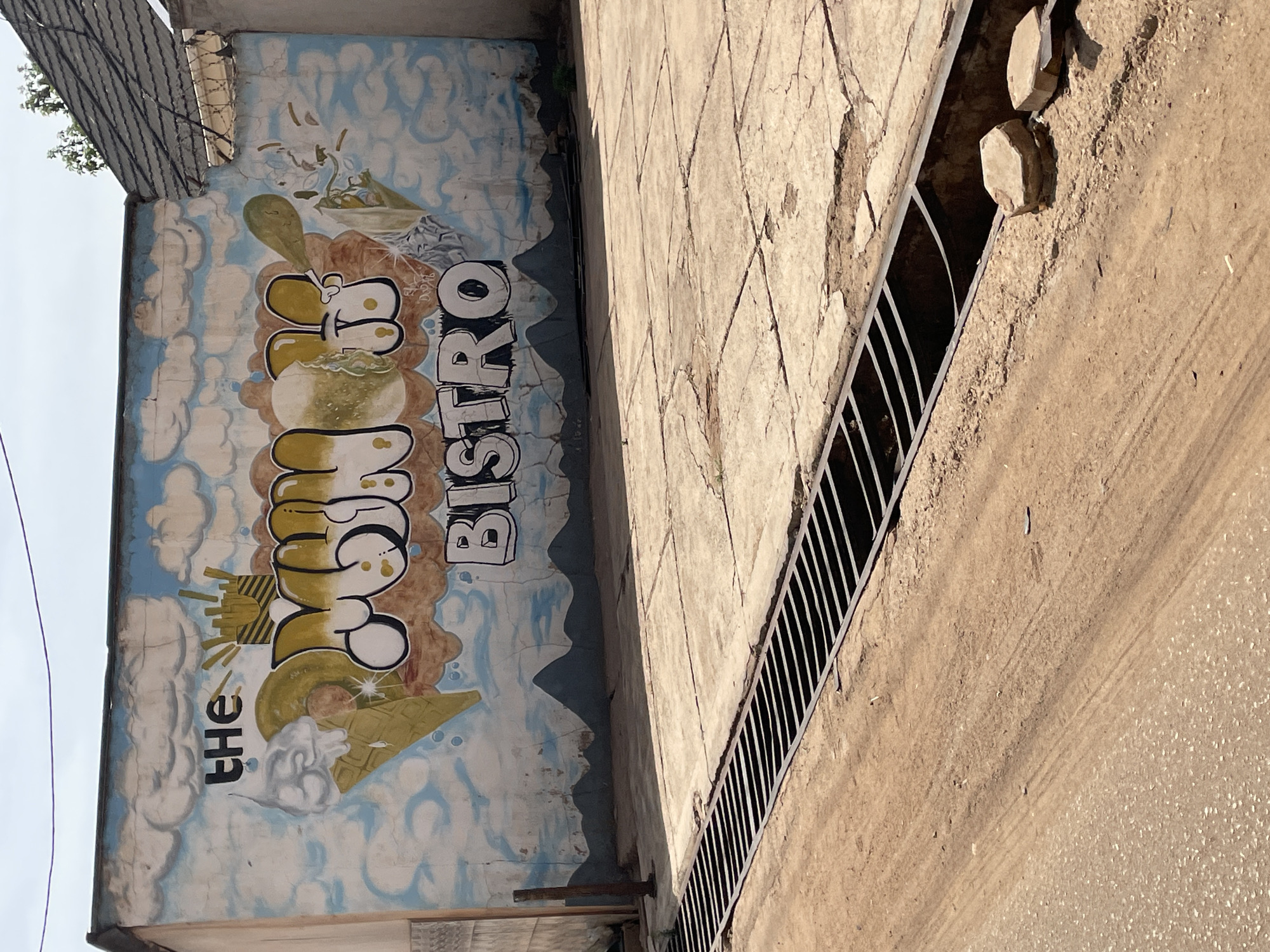 The image, rotated counter-clockwise, depicts a vibrant street art mural on a building wall, likely for a food establishment called "THE COOKIE BISTRO." The mural's background is a bright blue sky filled with white, puffy clouds. The text "THE COOKIE BISTRO" is painted in a distinctive graffiti style; "THE" is small, "COOKIE" is large and bubbly with yellow and brown tones, and "BISTRO" is rendered in white block letters with black outlines. Complementing the text are graphic elements such as an abstract yellow sunburst or explosion, a sparkling waffle cone-like shape, and a bowl containing green sprouts or plants. A dark grey, undulating shape resembling distant hills or mountains sits at the bottom of the mural.
Adjacent to the mural, a textured, light-colored concrete wall displays a pattern of etched diamond shapes and some visible cracking. Below this wall, a long, dark metal grate covers a drainage channel. The ground beyond the grate consists of light brown dirt and gravel, showing faint tire tracks. Two hexagonal-shaped stone blocks are placed on the ground near the grate.
In the top left of the frame, a segment of a corrugated metal roof with barbed wire is visible, suggesting an adjoining structure. Beyond this, a bright, clear sky and a small cluster of green foliage can be seen. The scene is bathed in bright daylight, indicating a clear, sunny day in Jos, Nigeria. No individuals or specific activities are present, emphasizing the urban landscape and the colorful artistic advertisement.