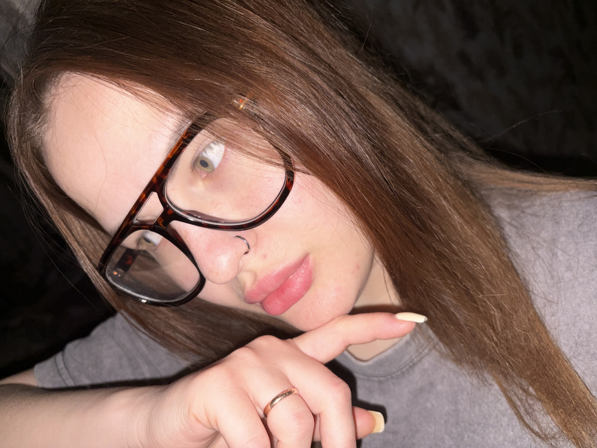 A close-up photograph depicts a young woman with light skin, wearing eyeglasses with brown frames. She has brown hair and a nose ring in her left nostril. Her lips are slightly parted, and she rests her hand on her cheek, a gold-colored ring visible on her finger. She wears a gray t-shirt. The background is dark and nondescript. The location is unknown and cannot be determined from the image.