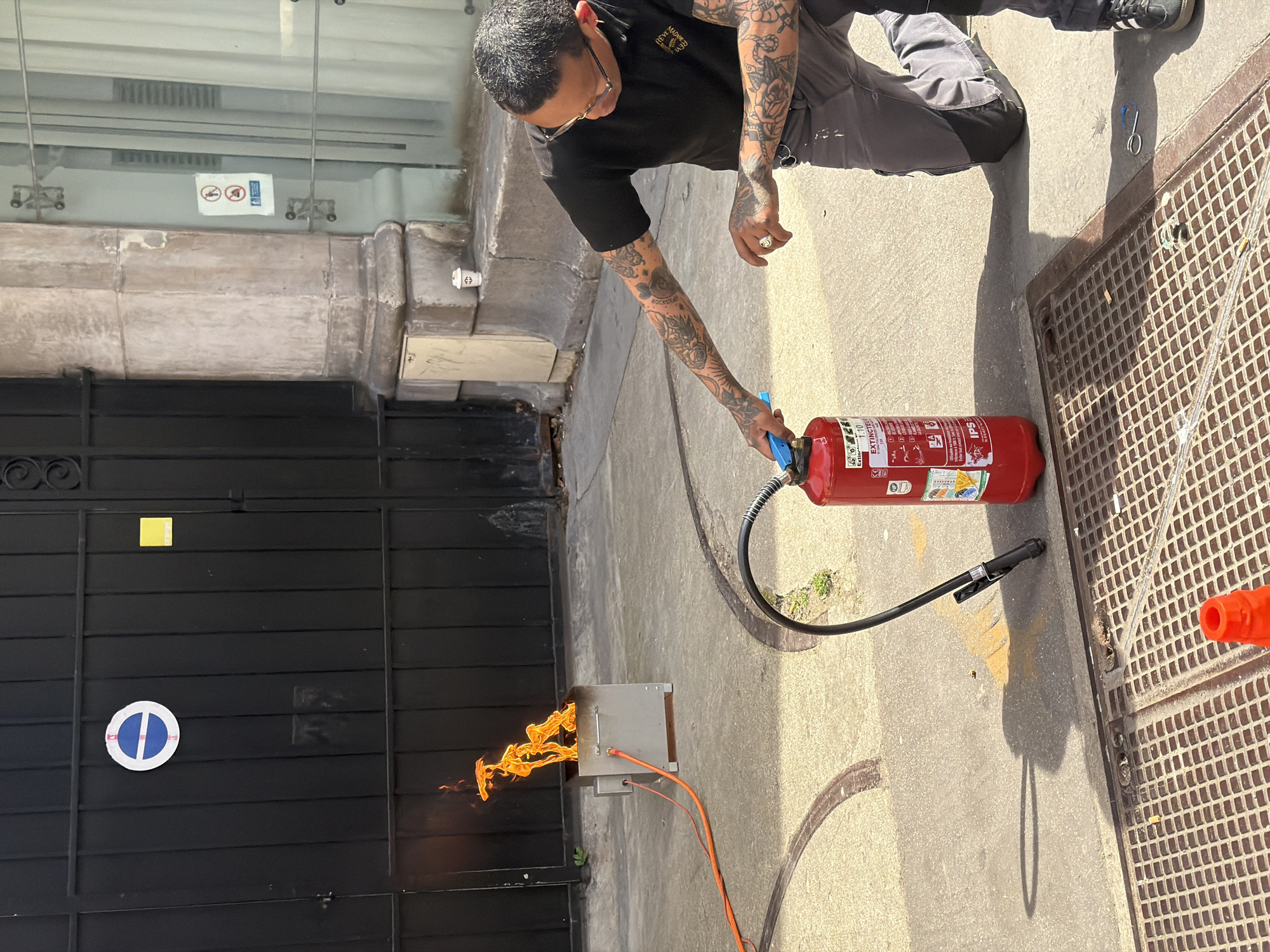 A person is seen handling a red fire extinguisher in an outdoor setting. Flames are emanating from a metal object, presumably an electrical box, on the ground near a black metal gate. The person has tattoos on their arms and is wearing a black shirt and gray pants. The setting appears to be a sidewalk in Paris, France, as indicated by the presence of a "no parking" sign on the gate, which matches the design of such signs in Paris. The ground is made of pavement and includes a metal grate or drainage cover. There are signs and details on the building visible in the background.