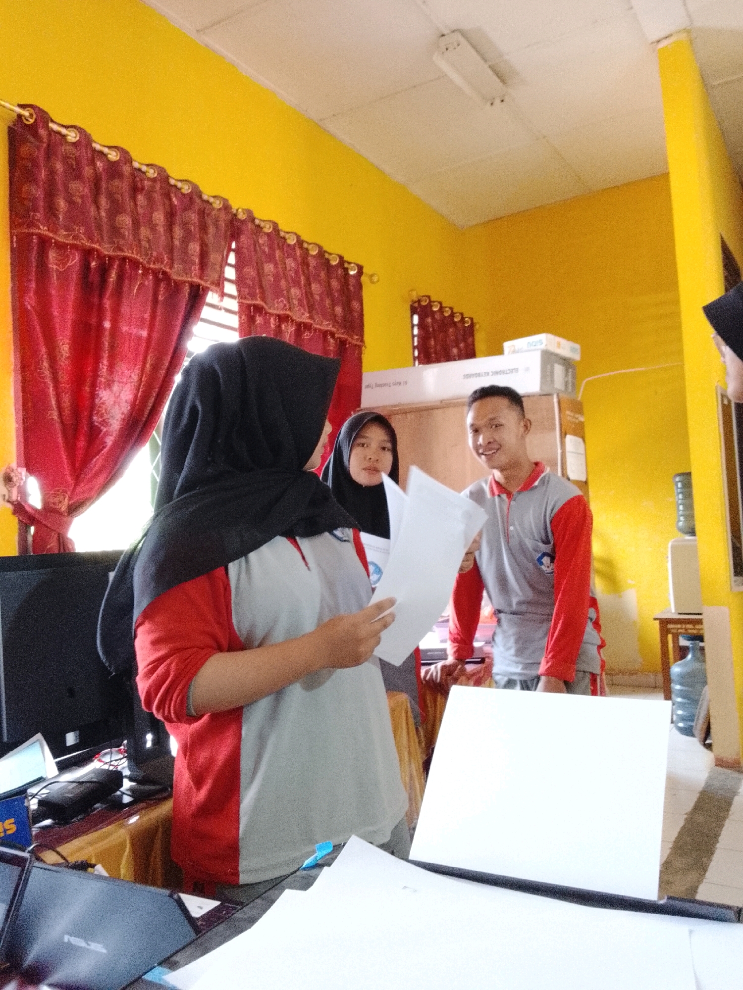 The image depicts an indoor scene with three individuals in what appears to be an office or classroom setting. The walls are painted bright yellow, and red curtains with gold accents hang on the left side of the frame, suggesting a decorative element. The ceiling is white with a fluorescent light fixture.
In the foreground, on the left, a person wearing a black hijab and a red and gray shirt is holding up white papers, looking towards the right. Their face is partially visible. To their right and slightly behind, another person, also wearing a black hijab and a dark garment, is visible from the shoulders up, looking towards the left.
In the middle ground, a young man in a red and gray polo shirt is smiling and looking directly at the camera while holding white papers. Behind him, stacked boxes are visible, one labeled with text that includes "ELECTRONIC KEYBOARDS". A water dispenser with a large blue jug is visible on a shelf to the right.
The immediate foreground shows a desk with laptops and papers, implying a workspace. The overall impression is a candid shot of people engaged in some sort of activity involving documents. There is no specific visual information in the image to definitively place the scene in Jepara, Indonesia.