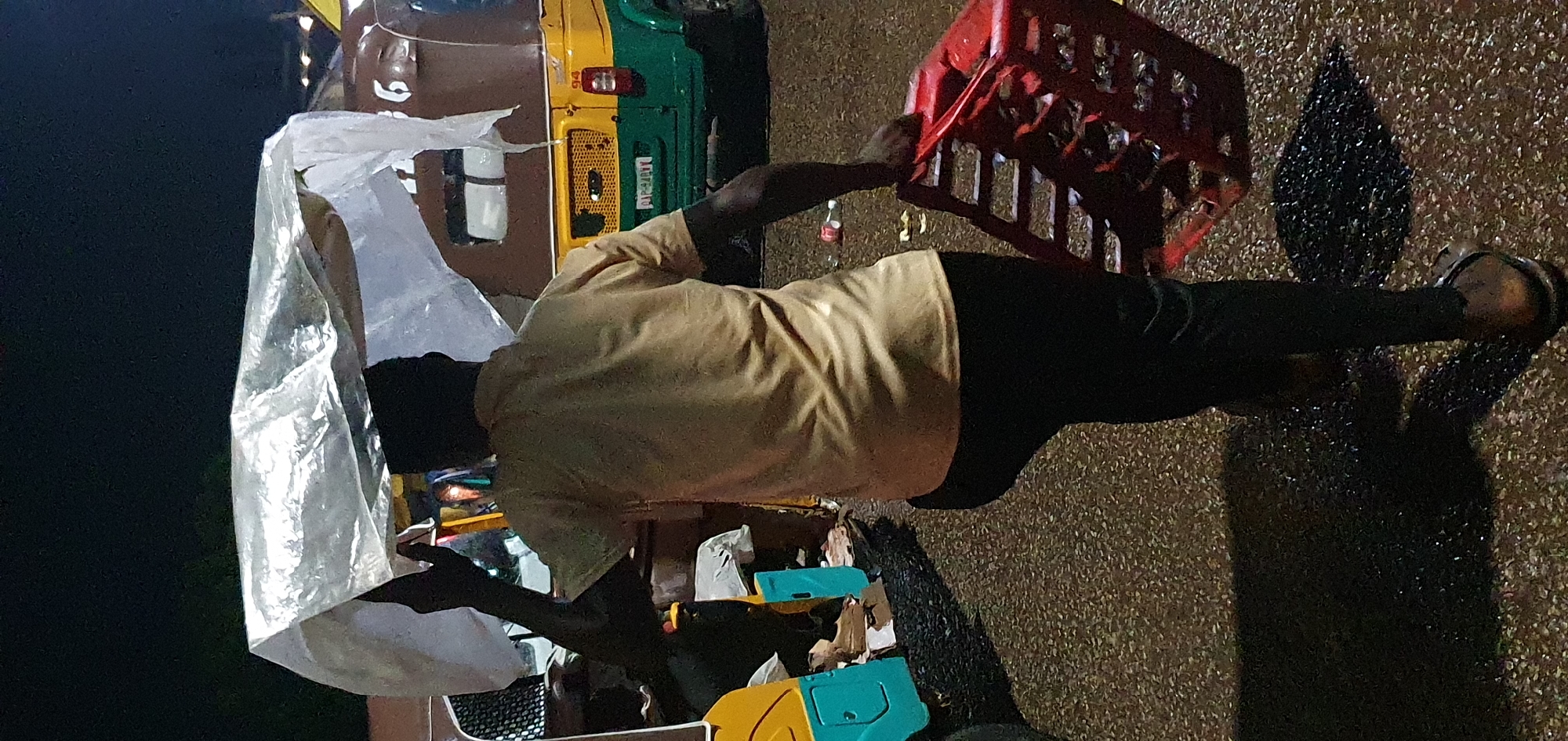 In the city of Bukuru, Nigeria, at night, a person walks away from the viewer, carrying a large, crinkled plastic sheet over their head as a makeshift umbrella. They are wearing a light-colored short-sleeved shirt and dark pants, and sandals. In their right hand, they are carrying a bright red plastic crate. Behind them, a brown, yellow, and green auto-rickshaw is parked, its headlights off. The ground is dark and appears wet, reflecting the ambient light from the street. The scene is dimly lit, suggesting it is nighttime.
