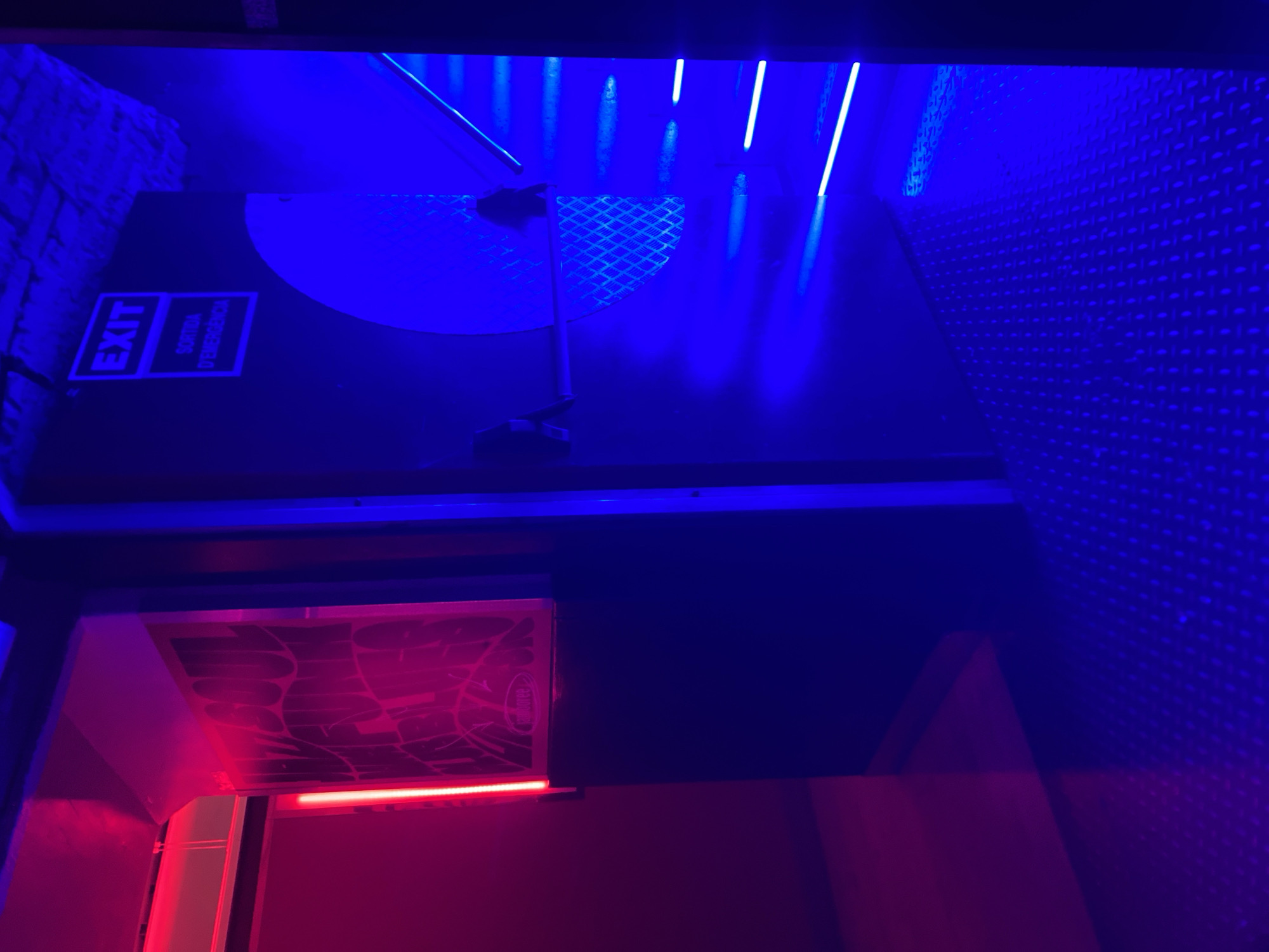 The image shows an interior scene illuminated by red and blue lights, possibly within a nightclub or entertainment venue in Barcelona, Spain. The left side of the image is dominated by a red glow, with a vertical neon light and a large graphic on a wall. The right side is illuminated by blue light, featuring an "EXIT" sign with "SORTIDA D'EMERGENCIA" below it. A metal staircase with blue neon lights along its edge is visible, along with a textured metal floor. The environment appears to be a passage or hallway leading to an exit. There are no people visible in the image.