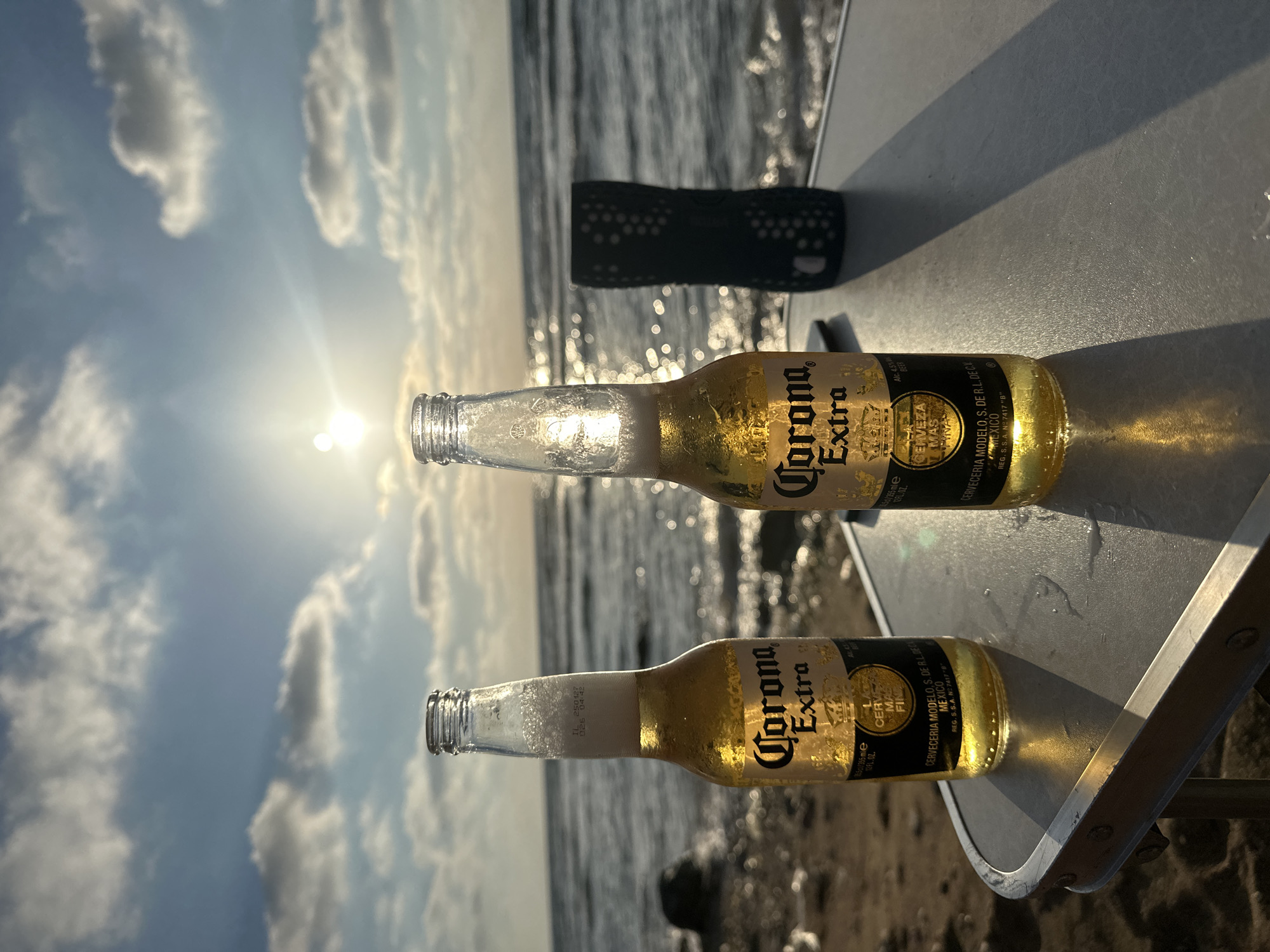 Two bottles of Corona Extra beer are placed on a folding table near the sea. The sun is setting, casting a warm glow over the scene. The bottles are condensation-covered, indicating they are cold. The background shows a calm sea with gentle waves and a sky filled with clouds illuminated by the setting sun. In the far right of the image, a dark, patterned cylindrical object, possibly a portable speaker, is visible. The ground in the foreground is sandy and appears to be part of a beach or a similar coastal area. The image conveys a sense of relaxation and leisure by the sea. The location is identified as the city of Haifa, Israel.