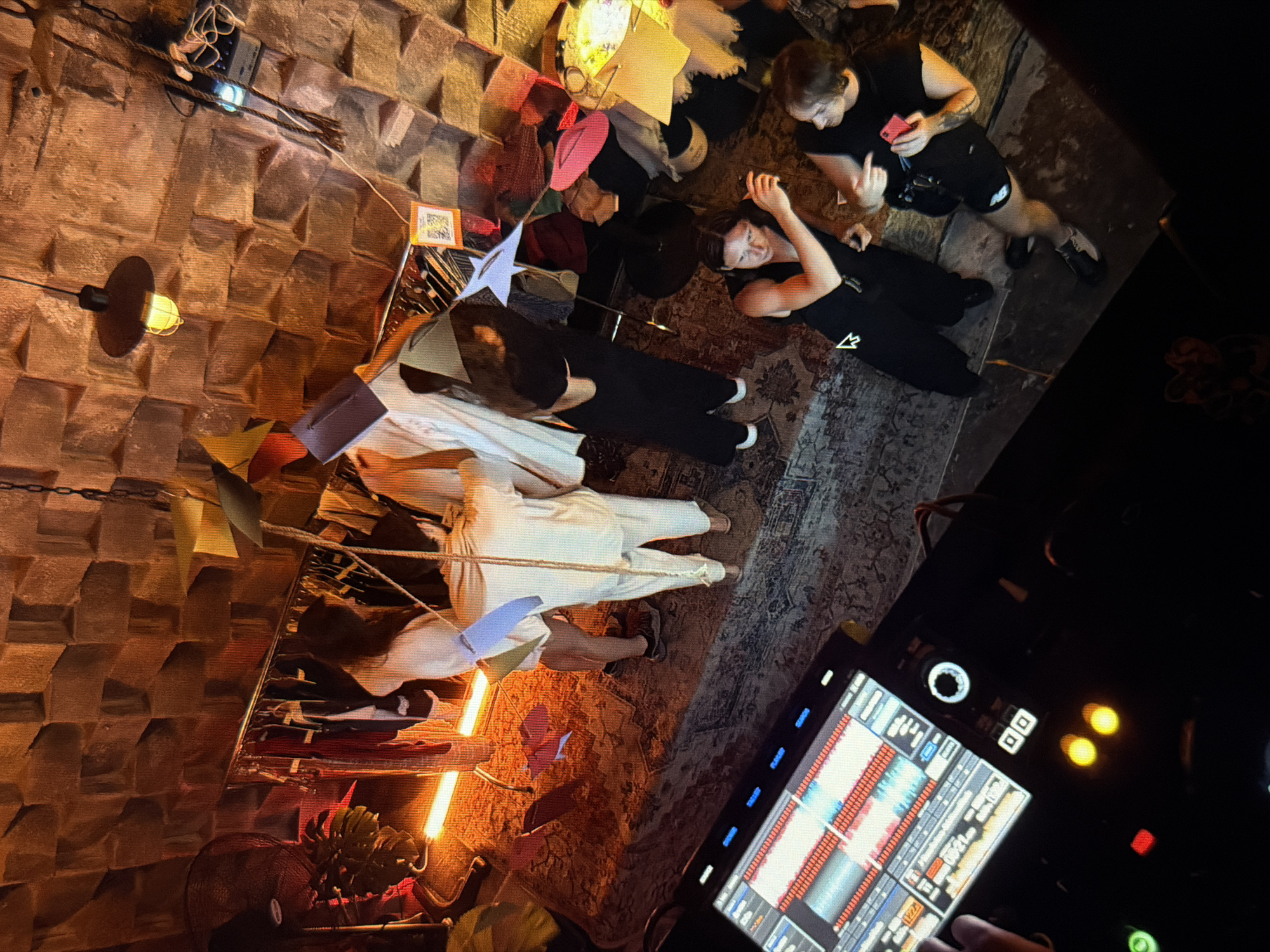 This indoor shot taken from above captures a gathering of several individuals in what appears to be a bar or lounge. The location is possibly in Đà Nẵng, Vietnam. Two individuals are situated in the upper right quadrant, one standing and looking at a smartphone while the other lies on the ground. Other people are arranged around what could be a table or display area, featuring colorful objects, papers, and decorations. The setting seems to be an event with people in various poses, some sitting and some standing. The lighting is dim and uses a mix of tungsten bulbs and neon, adding to the atmospheric feel. The walls have a unique textured design, which adds visual interest to the space. In the foreground, on the right side, is a DJ controller that may be used for playing music. The presence of the DJ controller and the overall atmosphere suggest an evening event or party.