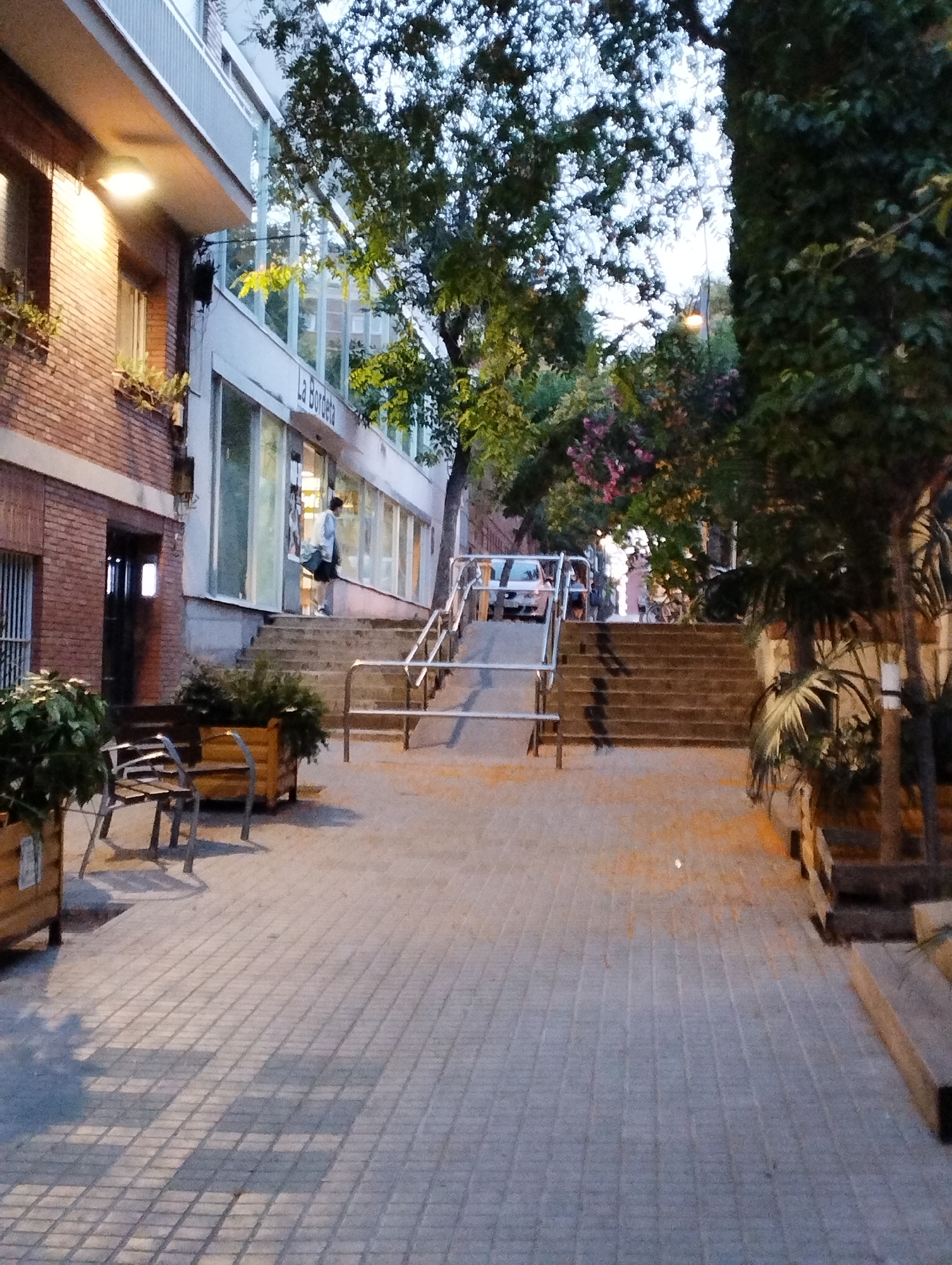 The image depicts a street scene in Barcelona, Spain, likely in the evening or early morning, as indicated by the artificial lighting and soft daylight. The foreground is a paved walkway with a brick pattern, leading towards a set of stairs with metal railings. To the left, a brick building with large windows is visible, with an illuminated sign that reads "La Bordeta." A person is standing in the doorway of the building, facing away from the camera. To the right of the stairs, there are several trees and plants, some with pinkish flowers. Further up the stairs, a light-colored car is parked. The overall atmosphere suggests a quiet urban setting with elements of greenery.