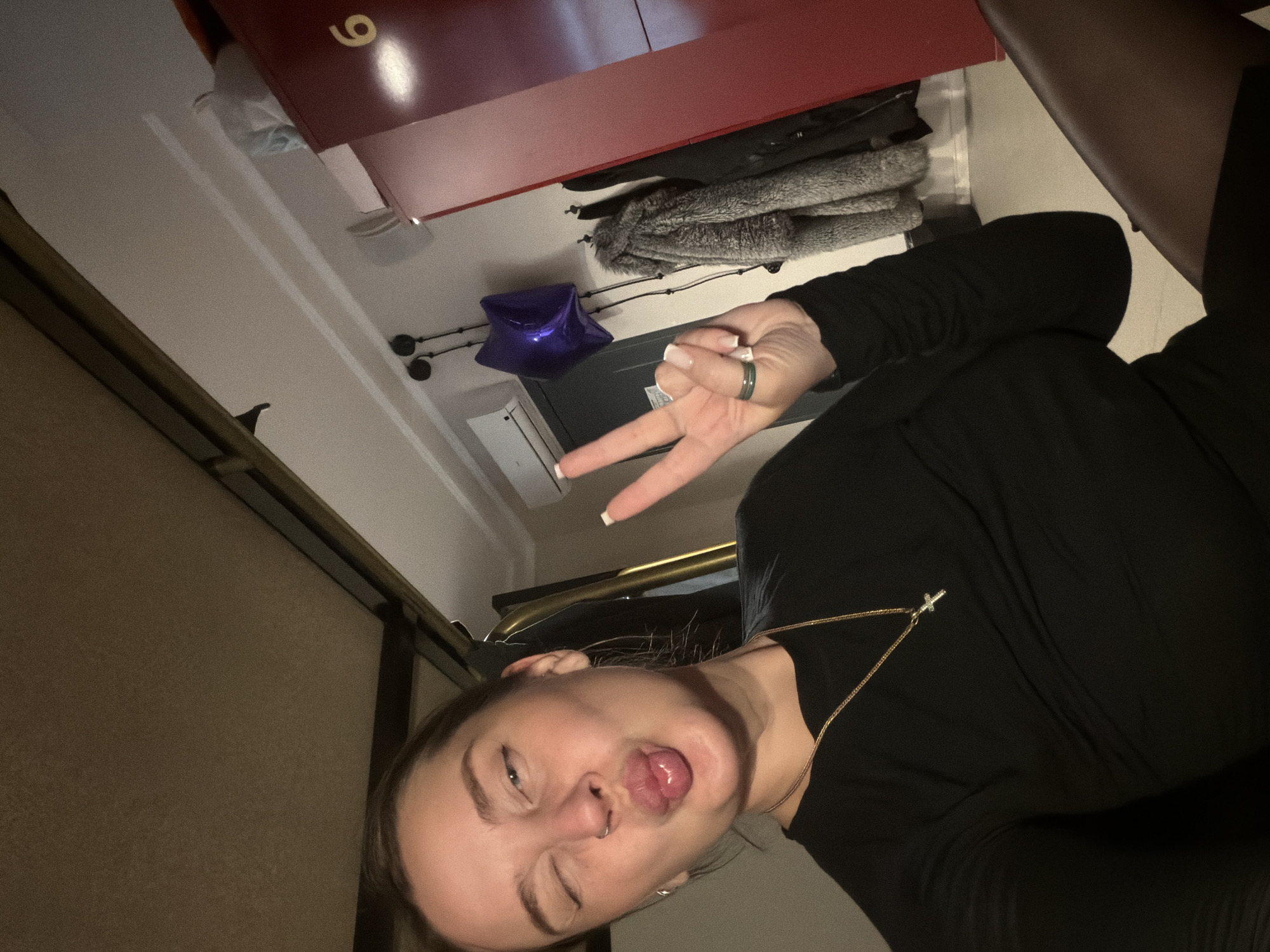 A woman poses indoors, sticking her tongue out and winking while giving a peace sign. She wears a black top, and a gold necklace with a cross is visible. A ring is on her finger. Behind her, a red cabinet with the number "9" is visible, along with clothes hanging on a rack and a blue star-shaped balloon. An air conditioning unit can be seen on the wall. The location is Moscow, Russia, based on the image context of the user and the type of environment.
