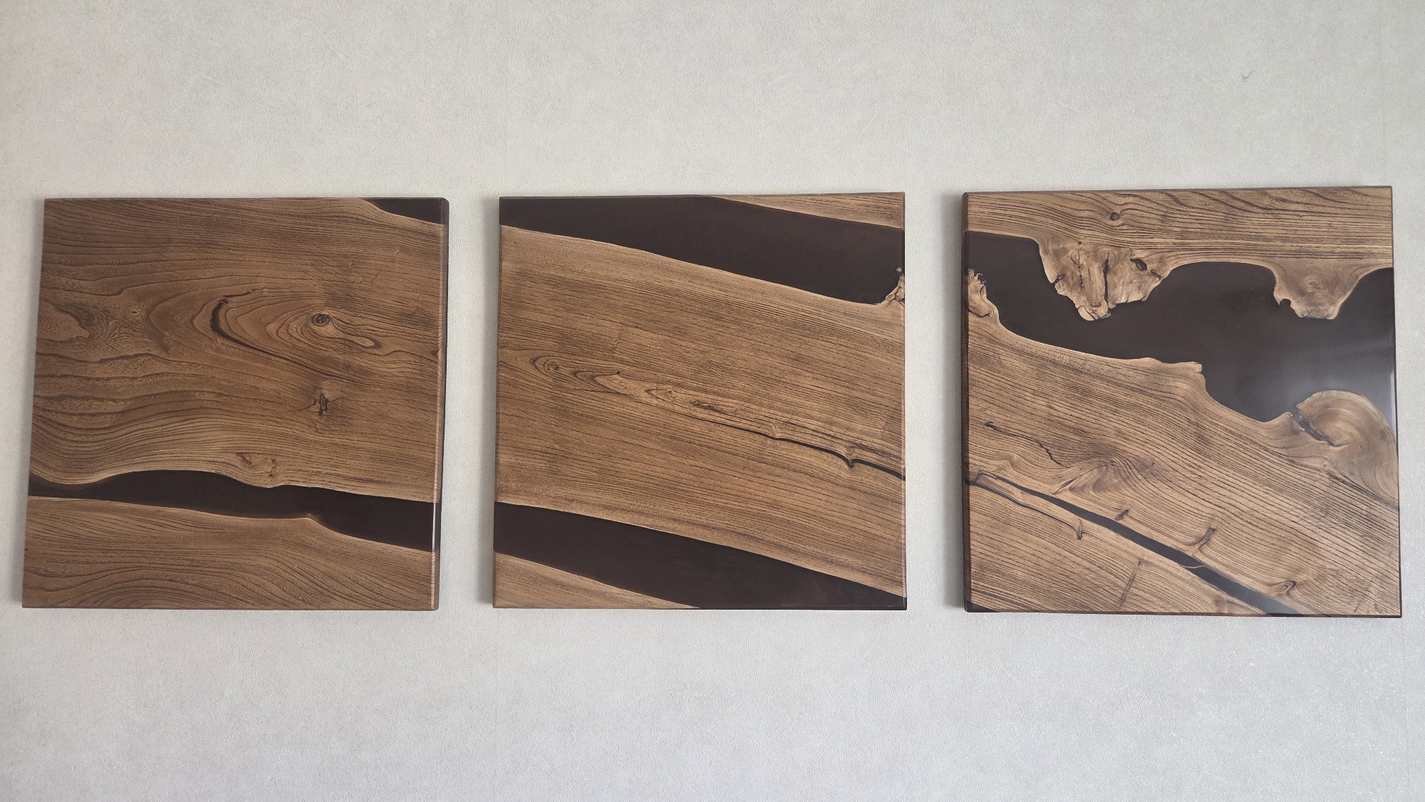 Three square wood and resin art panels are displayed against a light grey wall. The panels are arranged in a horizontal row. Each panel features light brown wood with a section filled with dark brown resin, creating a natural river-like effect. The wood grain is visible and varies in pattern across the three panels. The panels appear to be hung flush against the wall. The lighting is even and diffuse. No people are visible. There is no information present indicating that the photograph was taken in Chaikovsky, Russia.