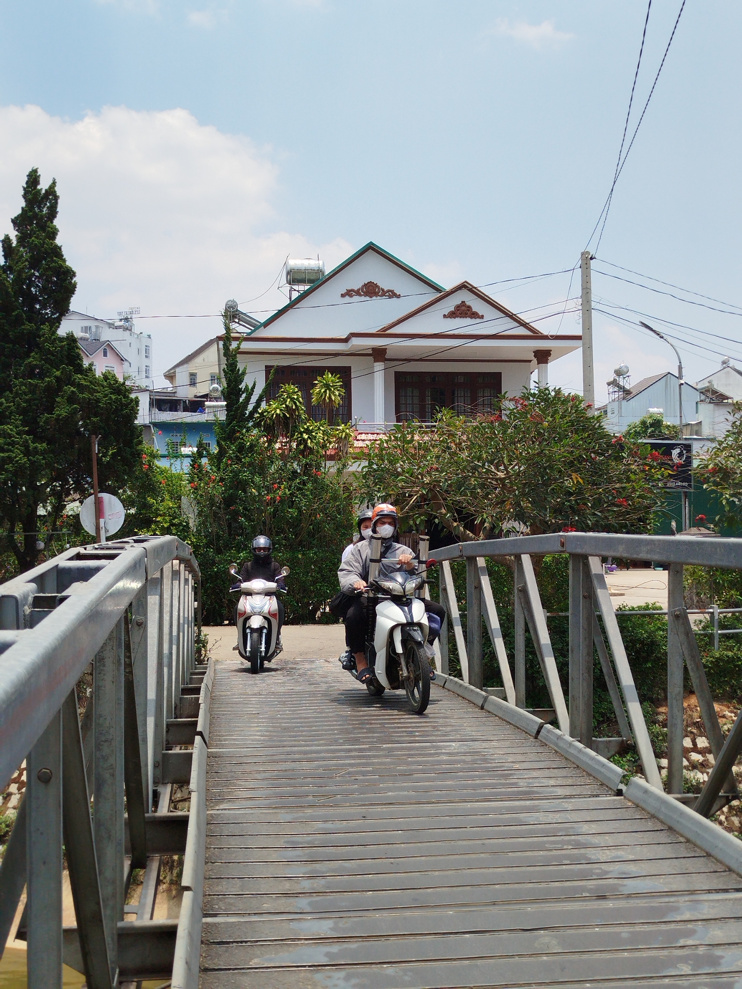 Two people on motorbikes are crossing a bridge. The bridge has a metal frame and a plank road surface. One person is wearing a grey helmet, while the other wears an orange helmet and a mask. The person with the orange helmet is riding a white motorbike with a black seat, while the person with the grey helmet is on a light silver motorbike. The background includes a white house with brown trim, lush greenery, and other buildings. Power lines are visible in the sky. The location is Cam Ly - Đà Lạt, Vietnam.