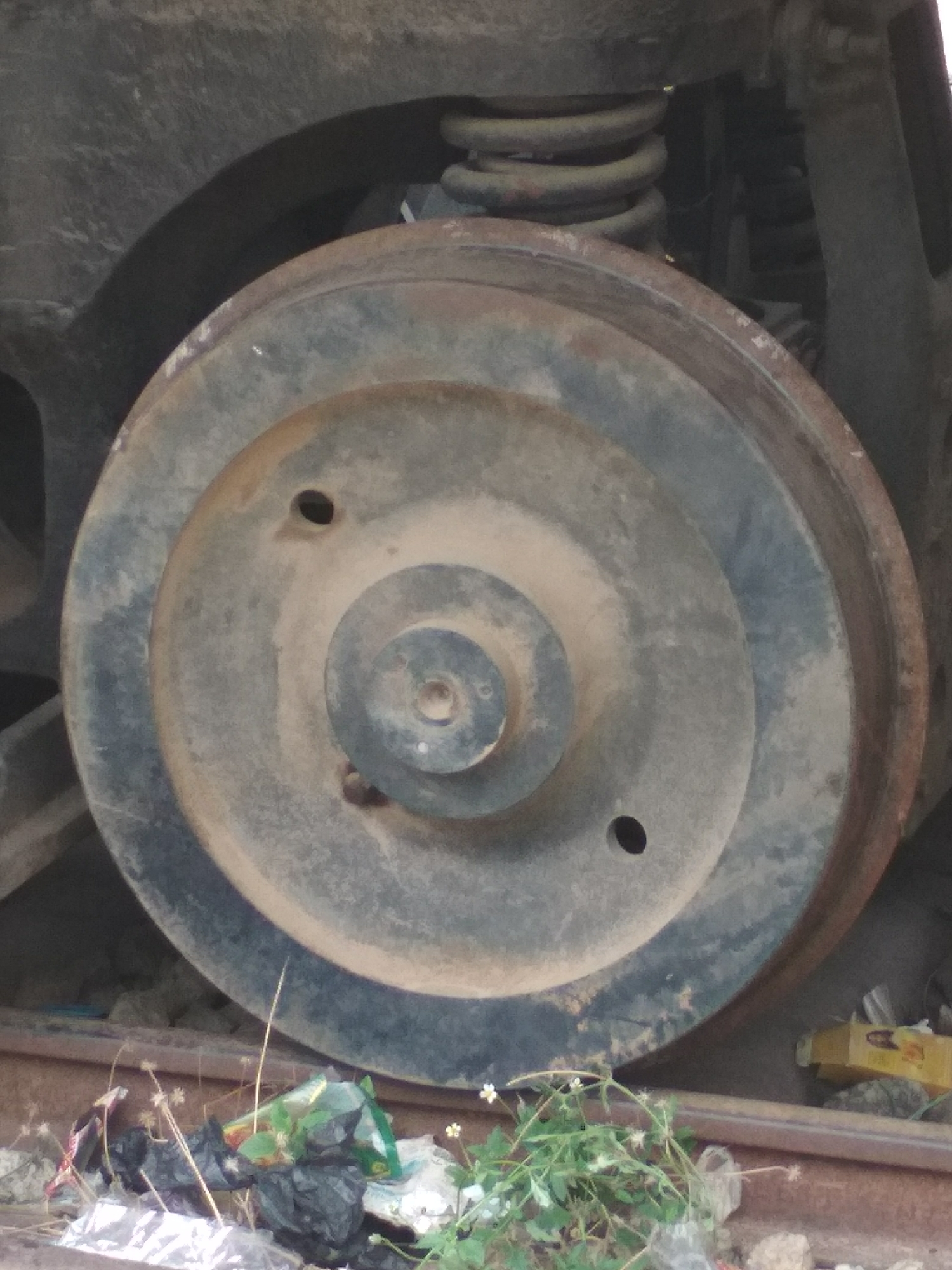 The image shows a large, dirty train wheel on a track in Jos, Nigeria. The wheel is steel and appears to be part of a railway car or locomotive. There are two prominent holes drilled into the wheel. The track beneath the wheel is partially obscured by trash, including plastic bags and other debris. The surrounding environment consists of gravel and vegetation. A spring and other parts of the train's undercarriage are visible behind the wheel.