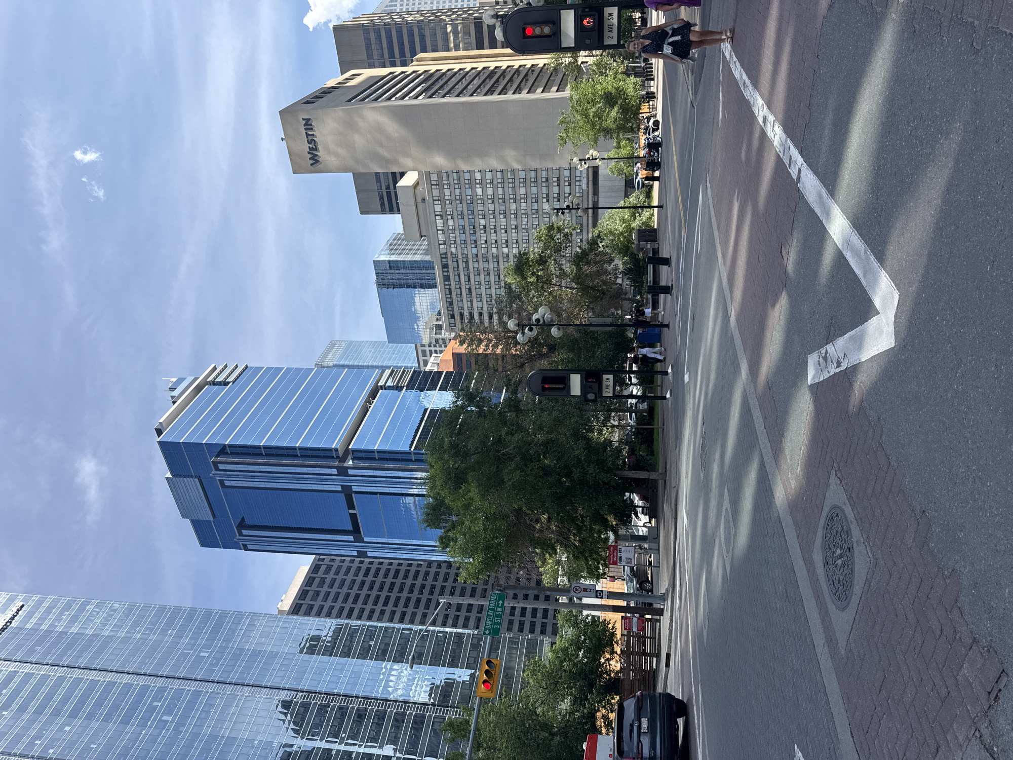This is a street-level, medium-shot photograph taken in downtown Calgary, Alberta, Canada, on a bright, partly cloudy day. The image captures an intersection with multiple modern high-rise buildings in the background. Dominating the left side of the frame is a tall, glass-facade skyscraper, reflecting the sky and surrounding structures. To its right, a shorter, deep blue glass-clad building stands out. Further back, a beige concrete building with "WESTIN" prominently displayed near its top is visible. The foreground shows a paved intersection with pedestrian crosswalks and road markings, including a large white checkmark painted on the asphalt. A few vehicles are present, including a dark SUV in the left foreground and other cars parked or driving in the mid-ground. Several trees with lush green foliage are interspersed between the buildings and along the sidewalk. A few pedestrians are seen walking on the sidewalks, one woman in a dark dress is in the right foreground. Traffic lights are visible, with one displaying a red light. Street signs indicating "BARCLAY PARADE" and "2 AVE SW" are also present. The overall scene depicts a bustling urban environment during daylight hours.
