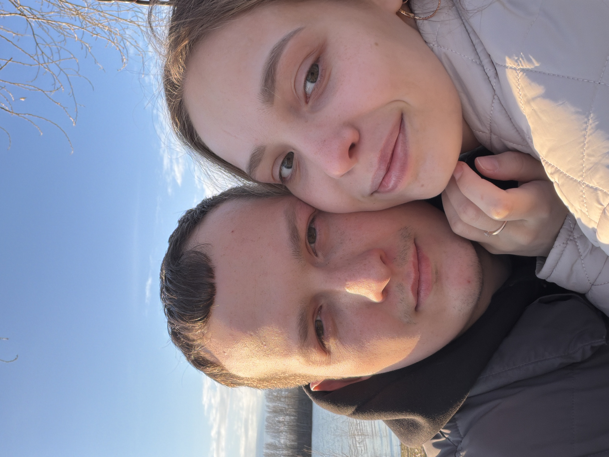 Two people are in the photo. A woman is leaning her head on a man's head. The woman is fair-skinned with brown hair, while the man has dark hair. The woman is wearing a cream-colored jacket and a gold earring. The man is wearing a gray jacket. In the background is a body of water and the sky. The photo was taken in the city of Chaikovsky, Russia.