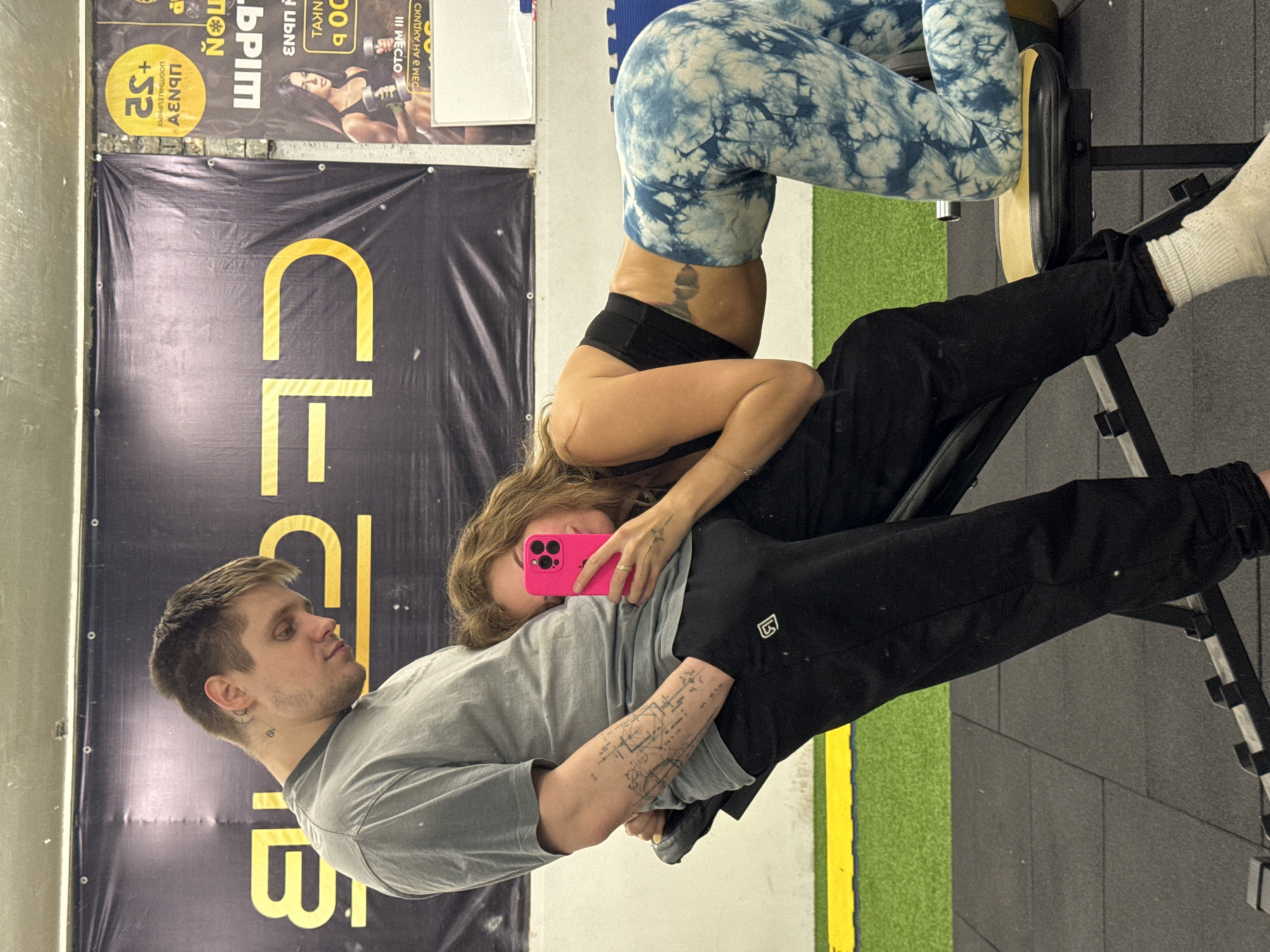 The image captures a playful or intimate moment between a man and a woman in what appears to be an indoor gym in Chaikovsky, Russia. The original image is oriented vertically, with subjects appearing sideways.
The woman is in an inverted position, resting her upper body on the lap or midsection of the seated man. Her legs are extended upwards, bent at the knees, with her feet braced against a black and tan padded gym bench or machine. She is wearing a black sports bra, black athletic shorts, and distinctive blue and white tie-dye patterned leggings. A tattoo is visible on her lower back/side. Her face is obscured by a bright pink smartphone she holds up, possibly taking a selfie or video.
The man is seated, wearing a grey t-shirt and black sweatpants. He has short brown hair, stubble, and notable tattoos on his left arm, featuring geometric or schematic designs, and a smaller tattoo on his neck. He looks upwards towards the woman with a neutral, attentive expression. His left arm is around or beneath the woman, while his right hand rests on his leg.
The background consists of a wall adorned with various gym banners and posters. A prominent black banner with large yellow letters displays "CLB," likely part of a gym name such as "CLUB." Other posters, featuring Cyrillic text, include "УКРЕПЛЕНИЕ" (strengthening) next to "+S2" on a yellow circular graphic, and fragmented phrases such as "III MECТO" (3rd place) alongside an image of a woman lifting weights. The flooring comprises dark grey interlocking gym mats, with a section of green artificial turf and a yellow dividing line visible. The Cyrillic text reinforces the provided location context.